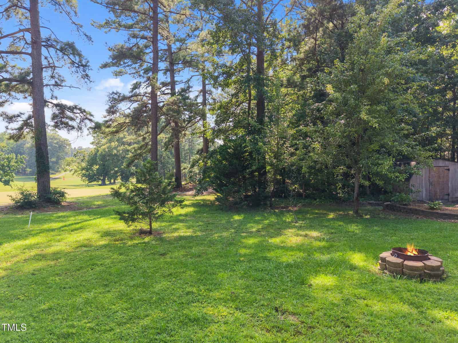 A grassy backyard with tall trees, a small fire pit made of stacked stones with a fire burning, and a wooded area in the background under a clear blue sky.