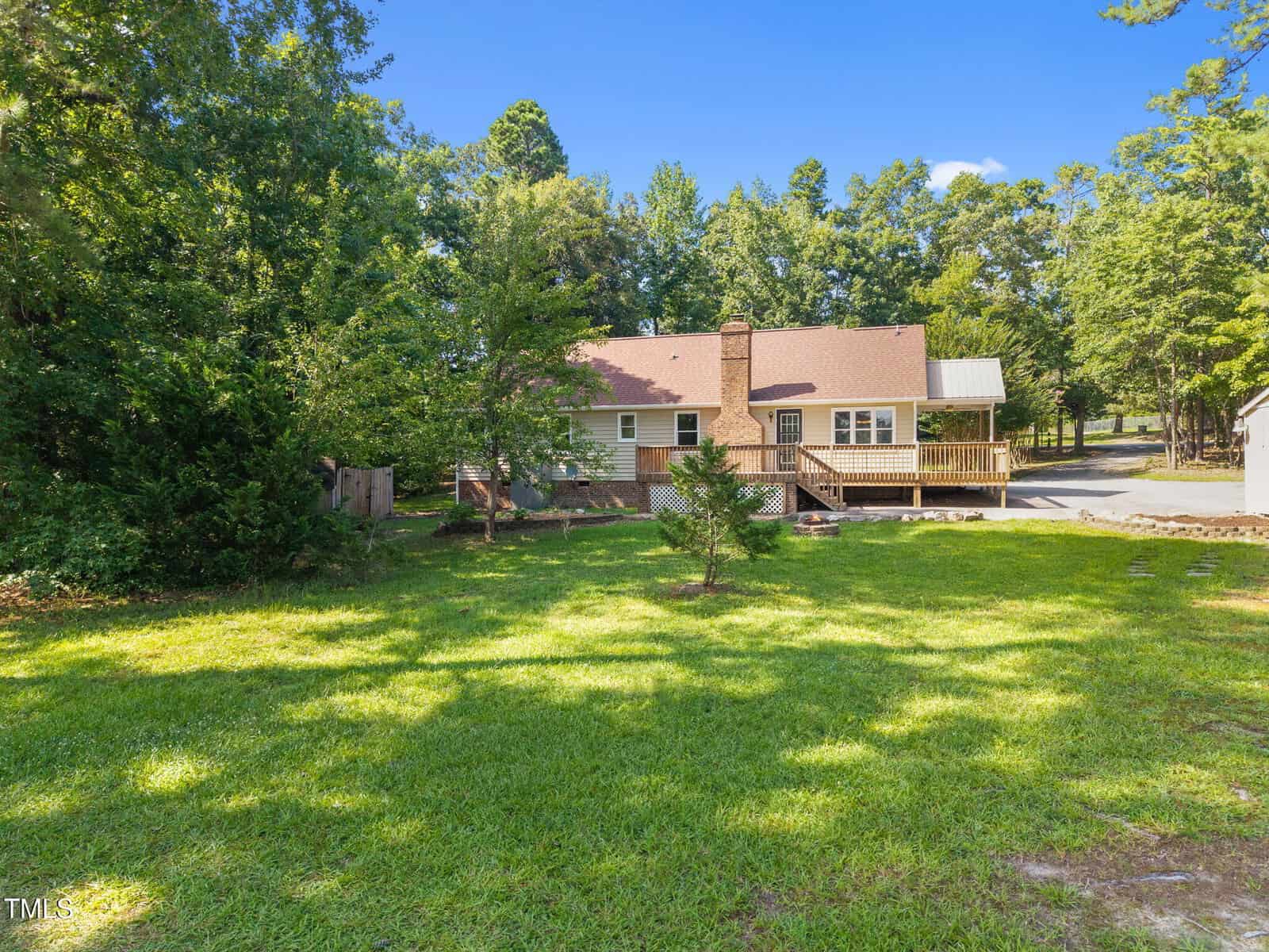 A tan house with a chimney and wooden deck sits among trees, facing a large, green grassy yard on a sunny day.