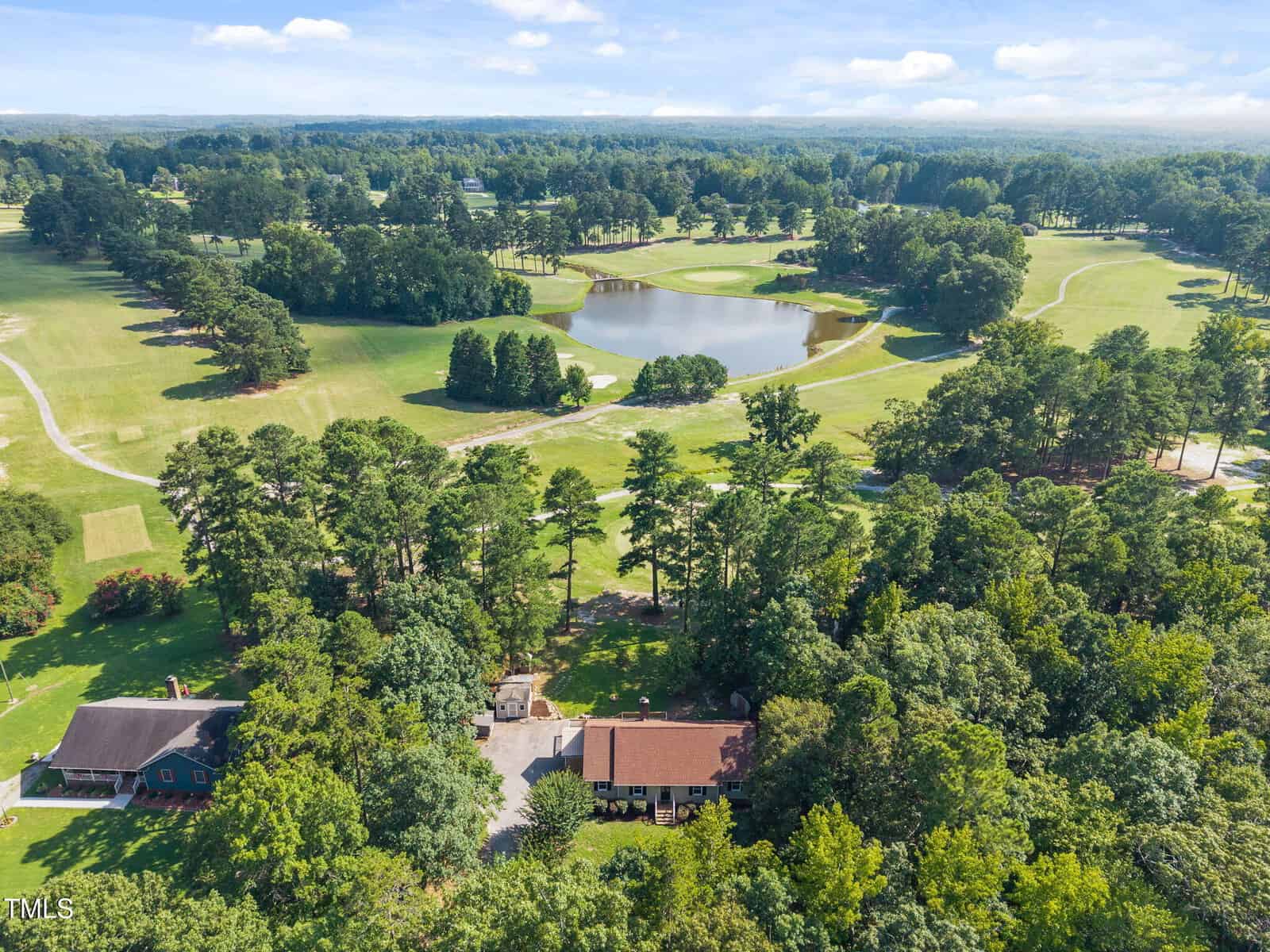 Aerial view of a house surrounded by dense trees, with a golf course, pond, fairways, and sand traps in the background under a blue sky with scattered clouds.