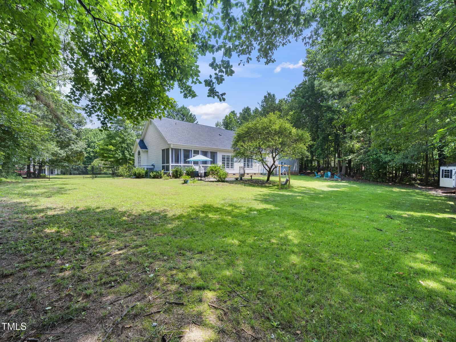A spacious backyard with green grass and trees, featuring a white house with a large patio, outdoor seating, and an umbrella on a sunny day.
