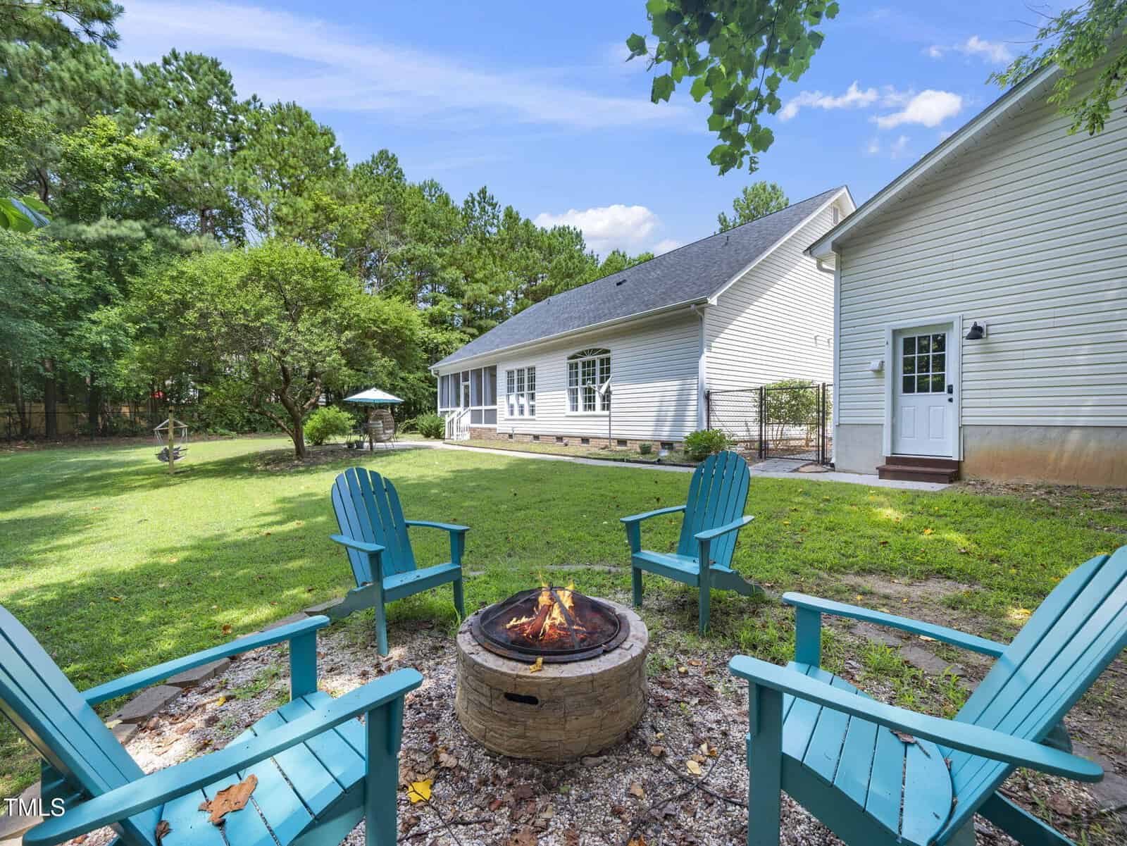 Four teal Adirondack chairs surround a small stone fire pit in a grassy backyard with a white house, trees, and a screened porch in the background under a blue sky with scattered clouds.
