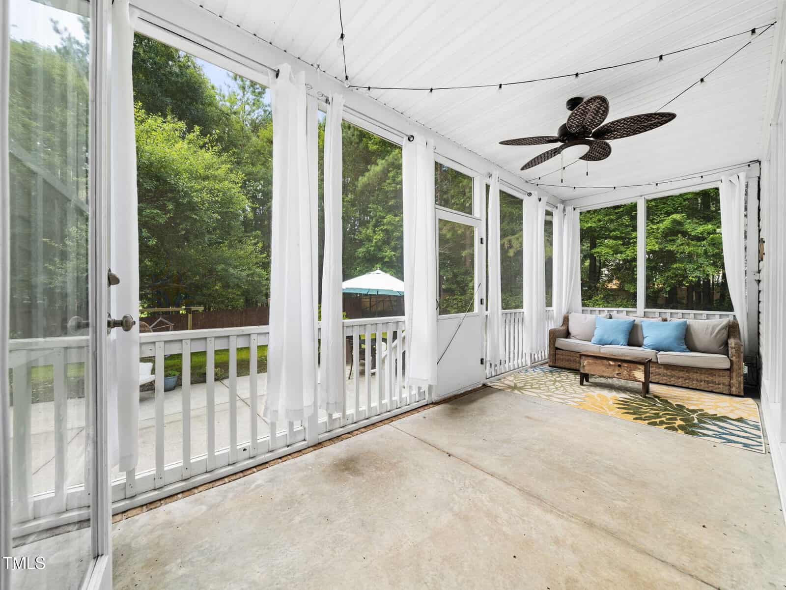 A screened porch with a ceiling fan, string lights, a wicker sofa with cushions, and a patterned rug. Large windows provide a view of a patio, yard, trees, and an outdoor umbrella. The floor is bare concrete.