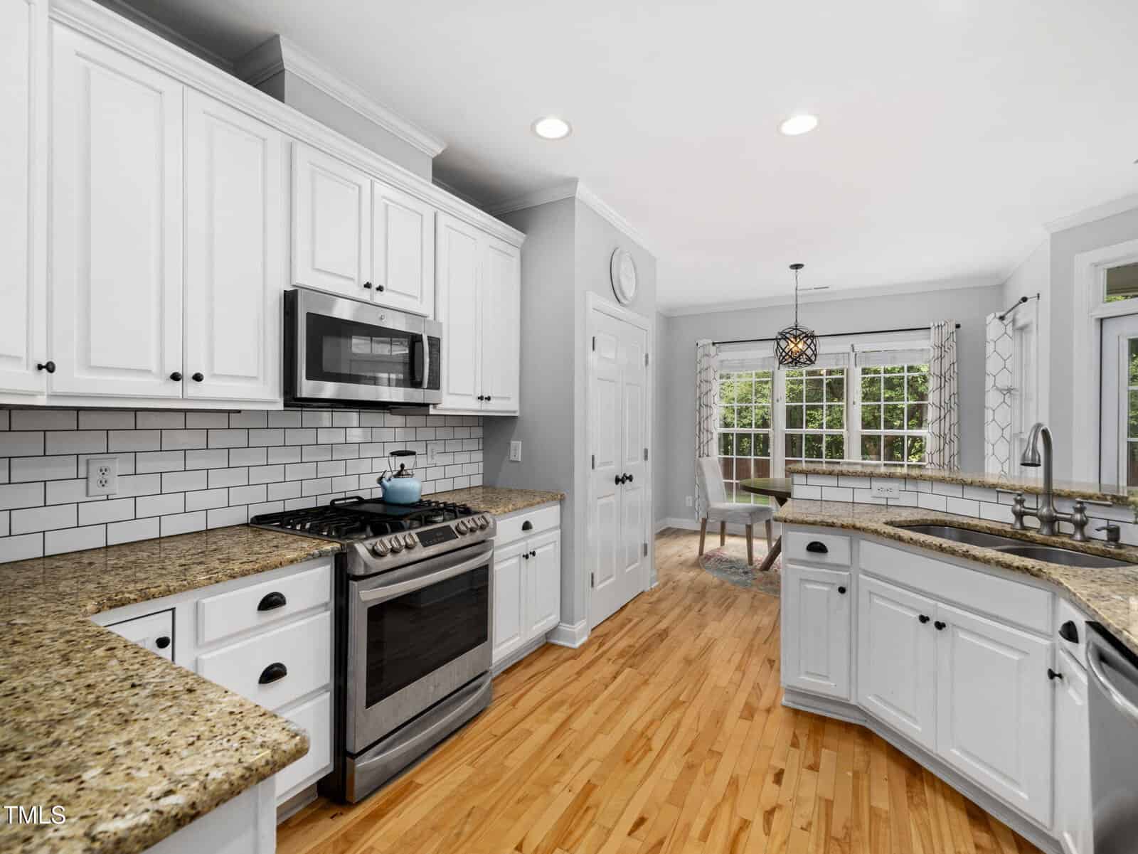 Modern kitchen with white cabinets, granite countertops, stainless steel appliances, subway tile backsplash, hardwood floors, and a breakfast nook with large windows and a hanging light fixture.