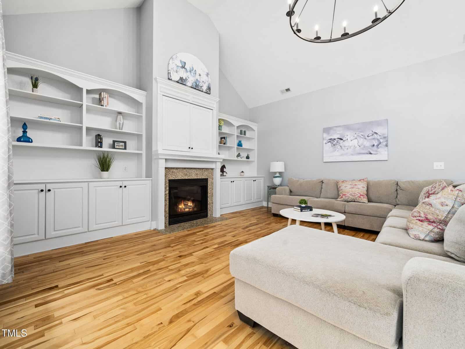 A bright living room with hardwood floors, a large beige sectional sofa, a round white coffee table, built-in white shelves, a fireplace with a mantel, and a modern chandelier on a high ceiling.