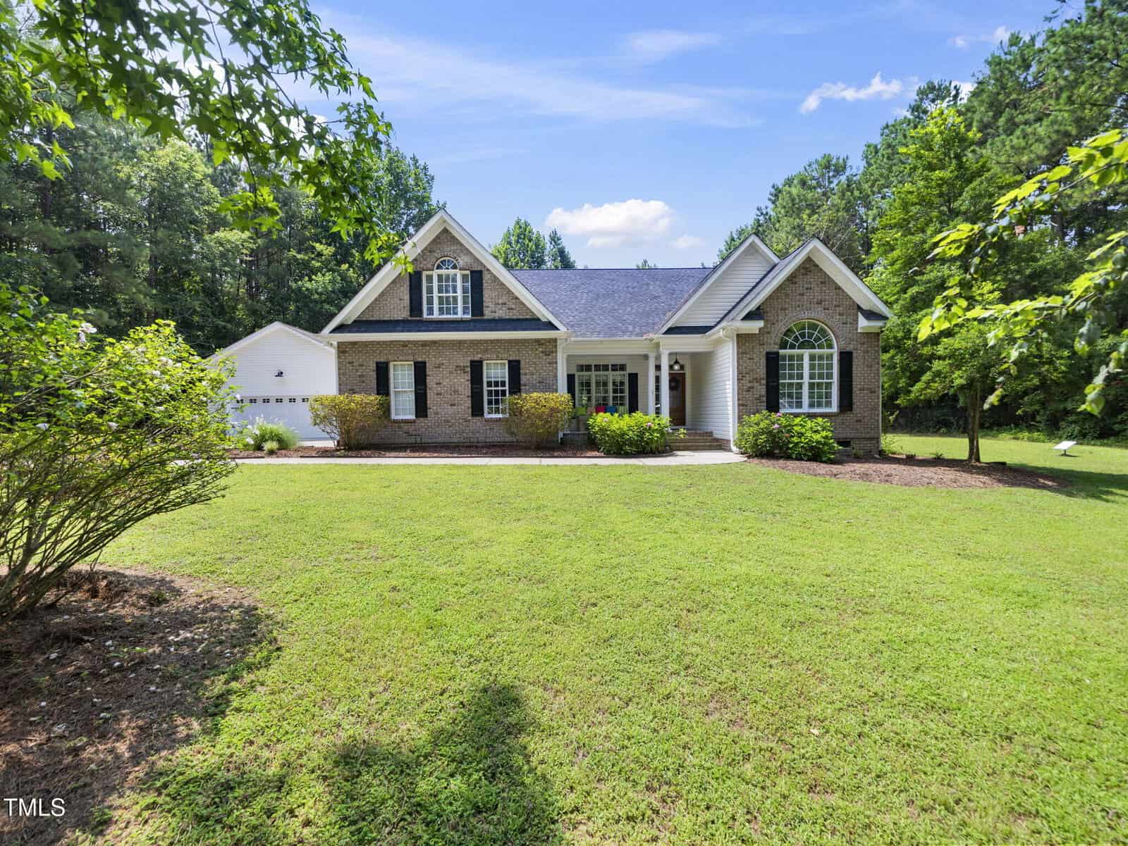 A single-story brick house with white trim and arched windows sits surrounded by green grass and trees under a blue sky with scattered clouds. A detached white garage is visible on the left.