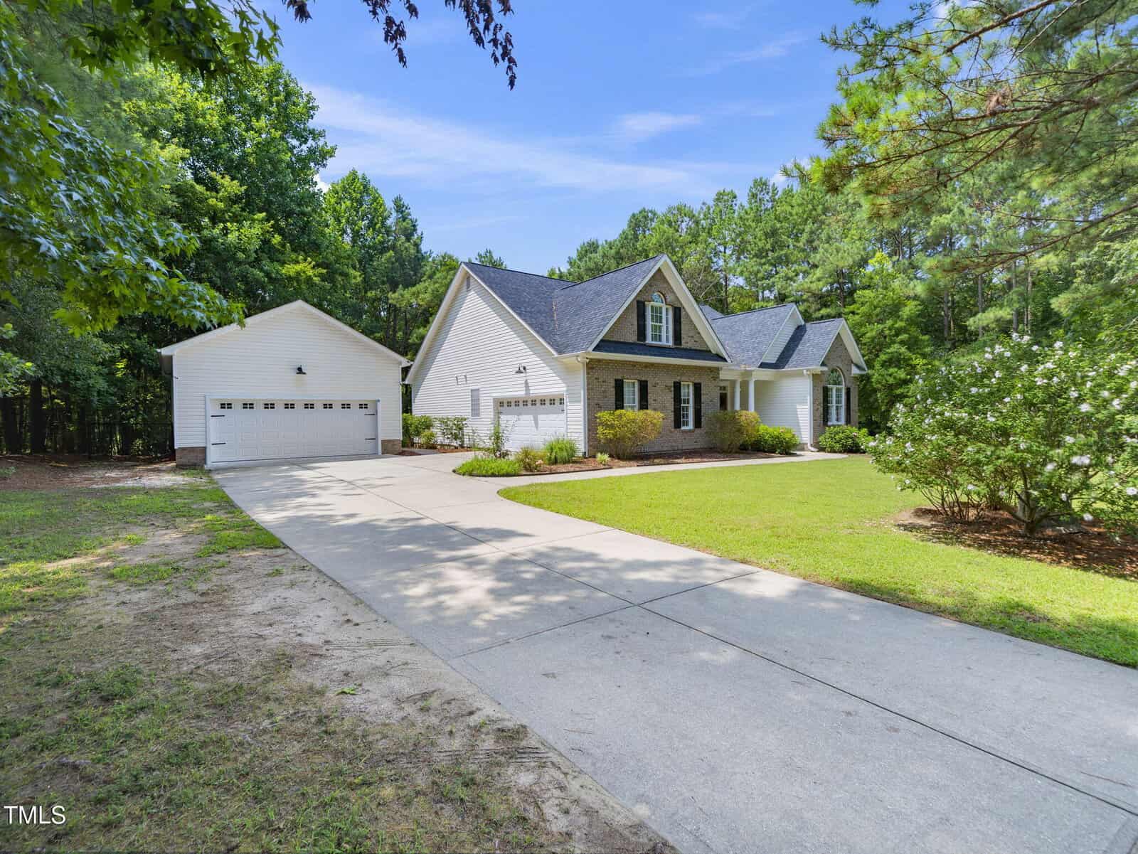 A white single-story house with a gray roof, attached two-car garage, and a long concrete driveway, surrounded by green grass, trees, and shrubs on a sunny day.