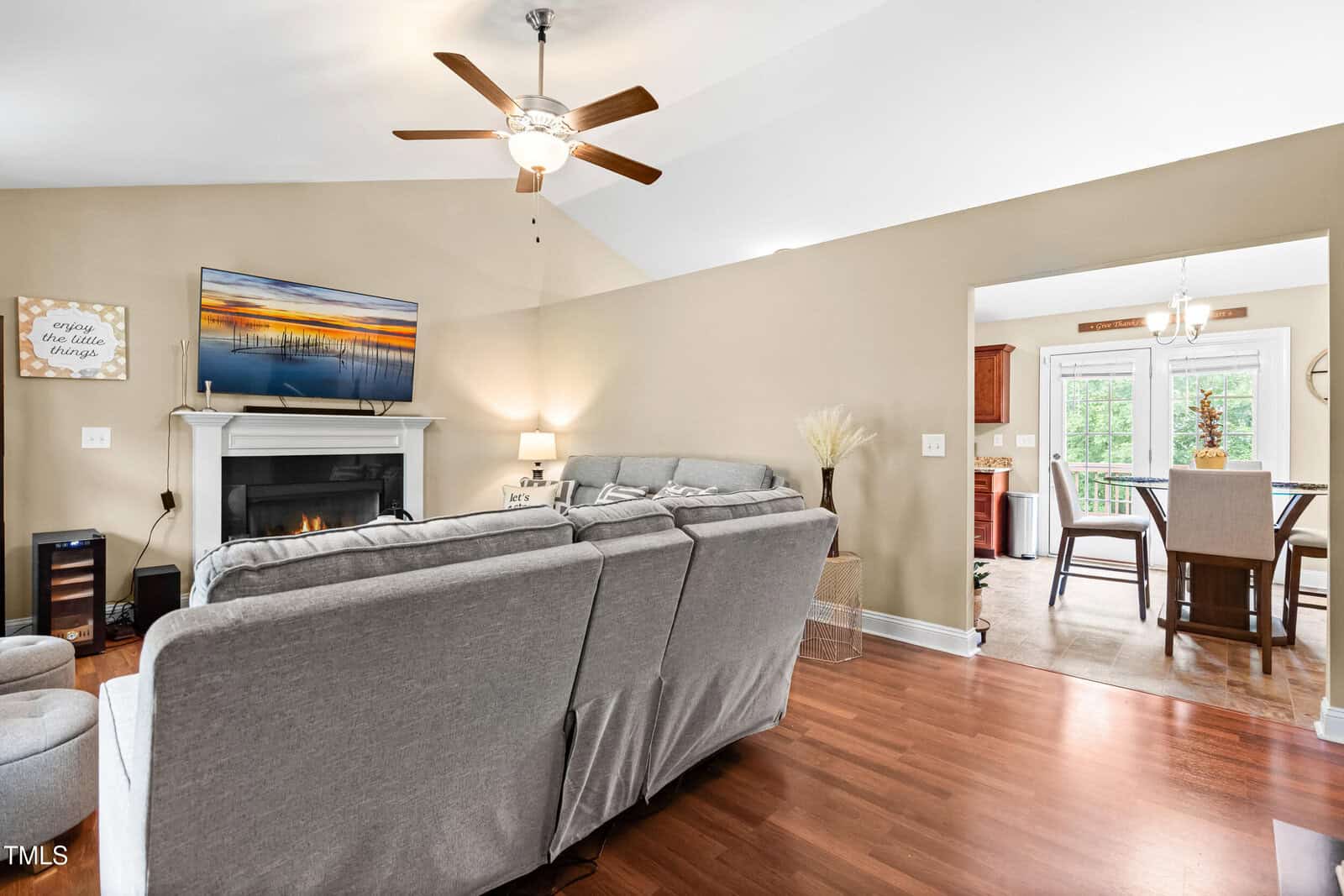 A living room with a gray sectional sofa facing a wall-mounted TV above a fireplace, a ceiling fan, wooden floors, and an open doorway leading to a dining area with a table and chairs.