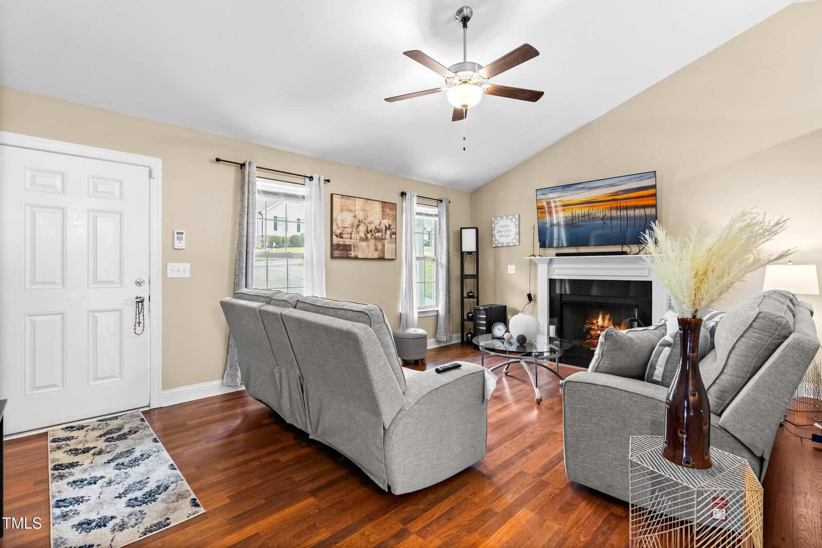 A living room with light gray sofas facing a fireplace and wall-mounted TV, hardwood floors, beige walls, and a ceiling fan. A white front door, windows with drapes, and decorative accents are also visible.