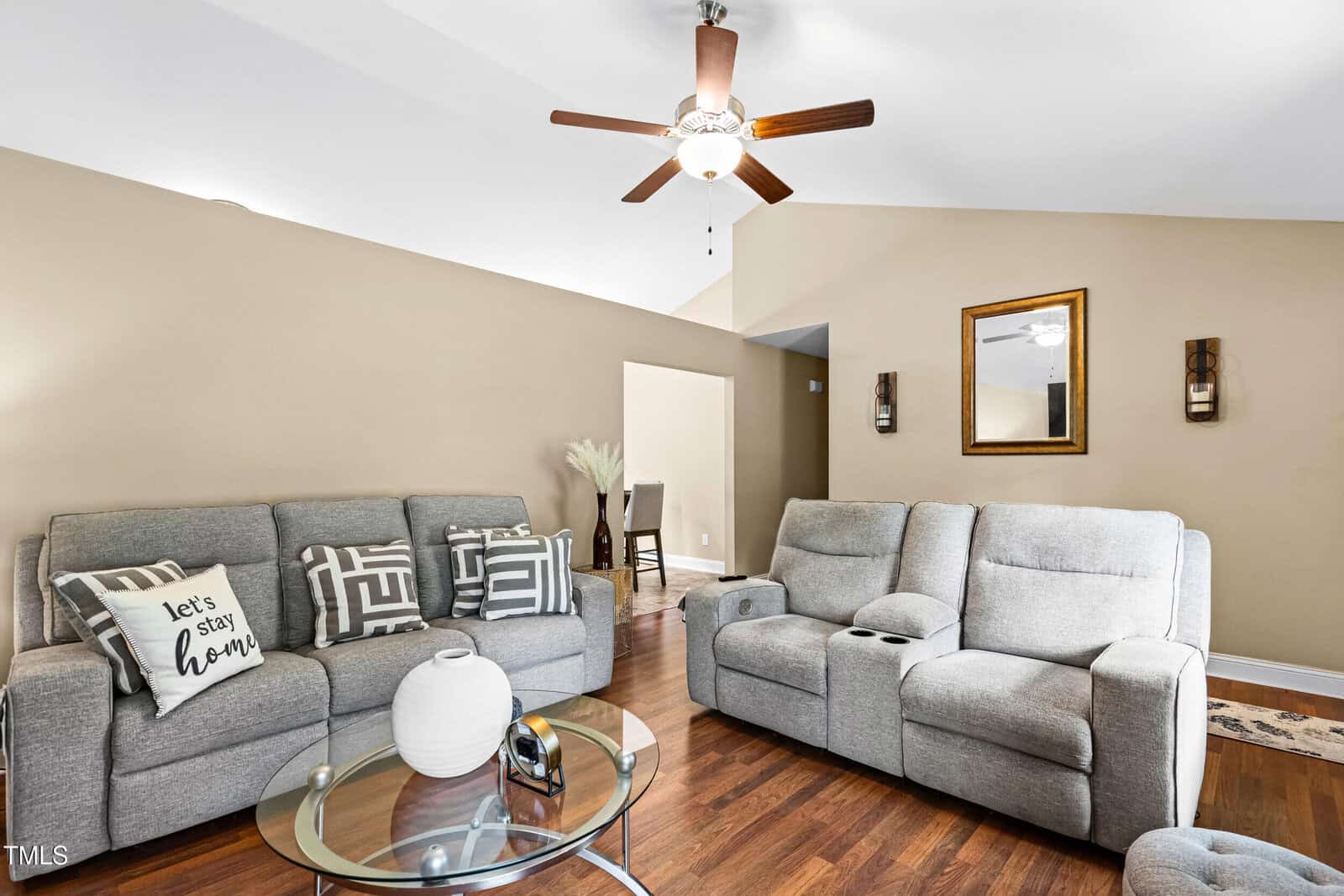 A living room with gray sofas, a glass coffee table with a white vase, wooden floors, beige walls, a ceiling fan, and wall decor including a mirror and two sconces. A pillow reads "Let's stay home.