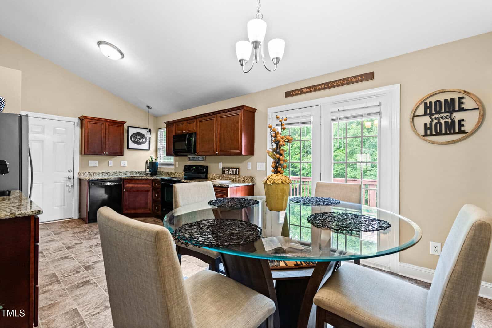 A kitchen with brown cabinets, granite countertops, and stainless steel appliances. A round glass dining table with four chairs is in the foreground, and a door leads to a balcony. Wall decor includes home-themed signs.