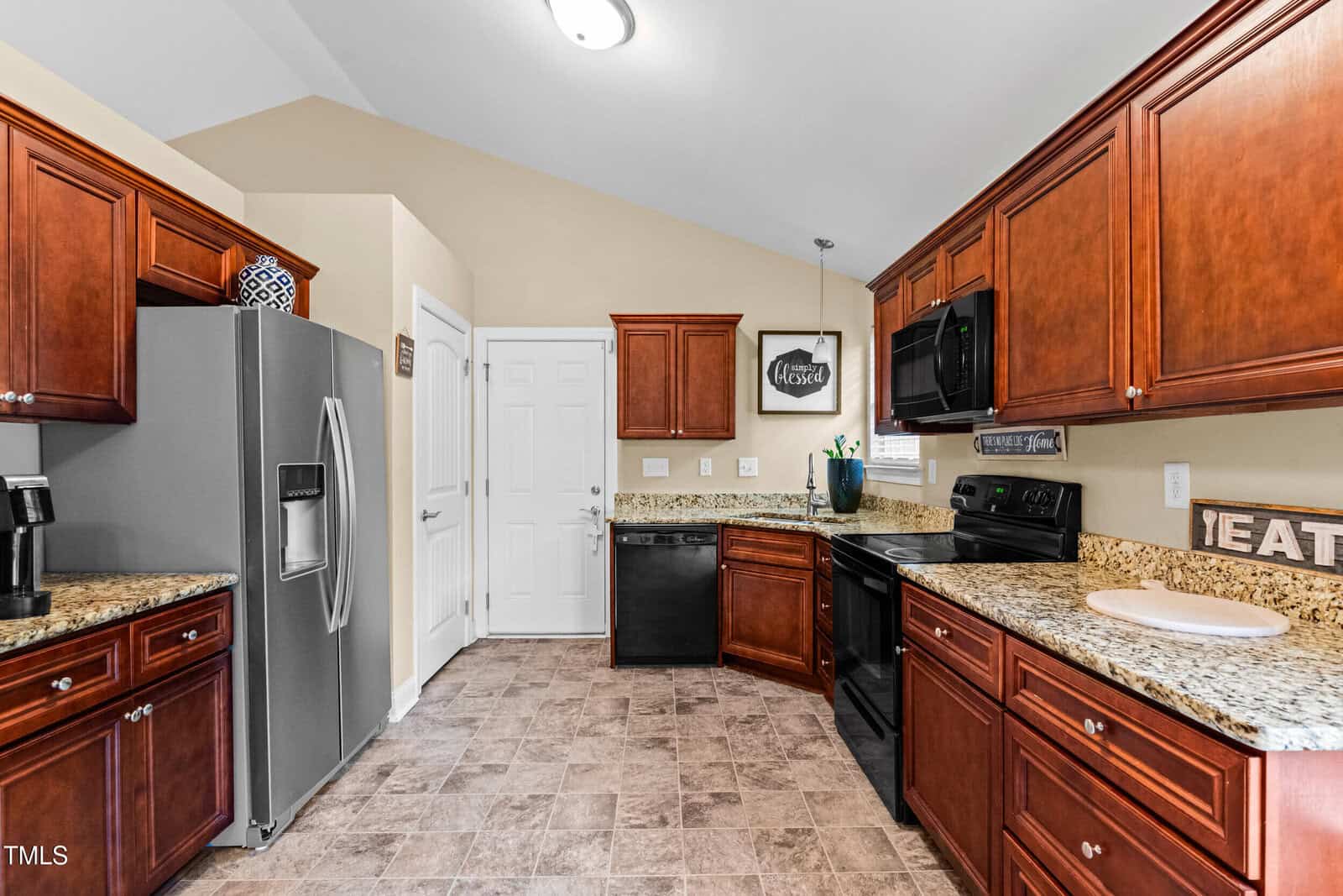 Kitchen with brown wooden cabinets, granite countertops, stainless steel refrigerator, black stove and dishwasher, beige walls, tile flooring, and decorative signs on the walls and counters.