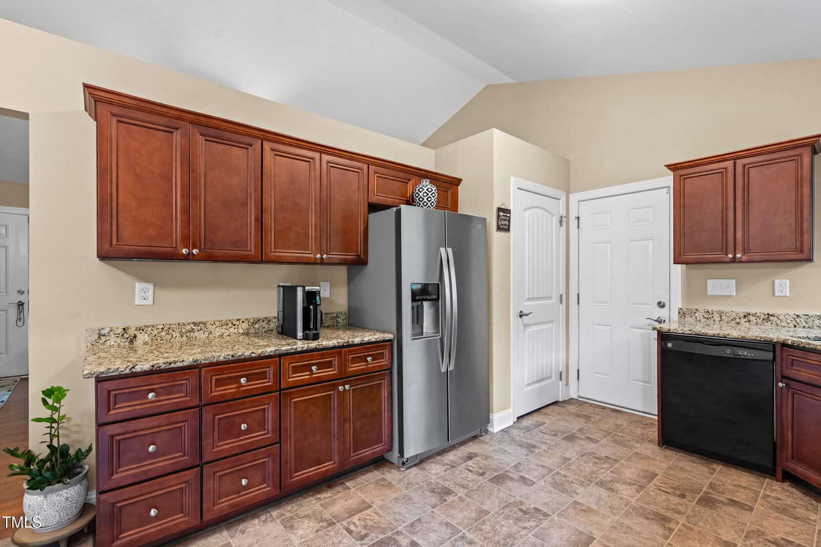 A kitchen with brown wood cabinets, granite countertops, a stainless steel refrigerator, a black dishwasher, a coffee maker, tiled flooring, and a potted plant in the corner.