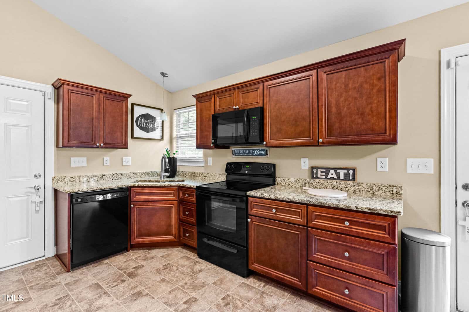 A kitchen with brown wooden cabinets, granite countertops, black appliances including a stove and dishwasher, beige walls, tile flooring, and a window above the sink. A decorative sign reads "EAT.
