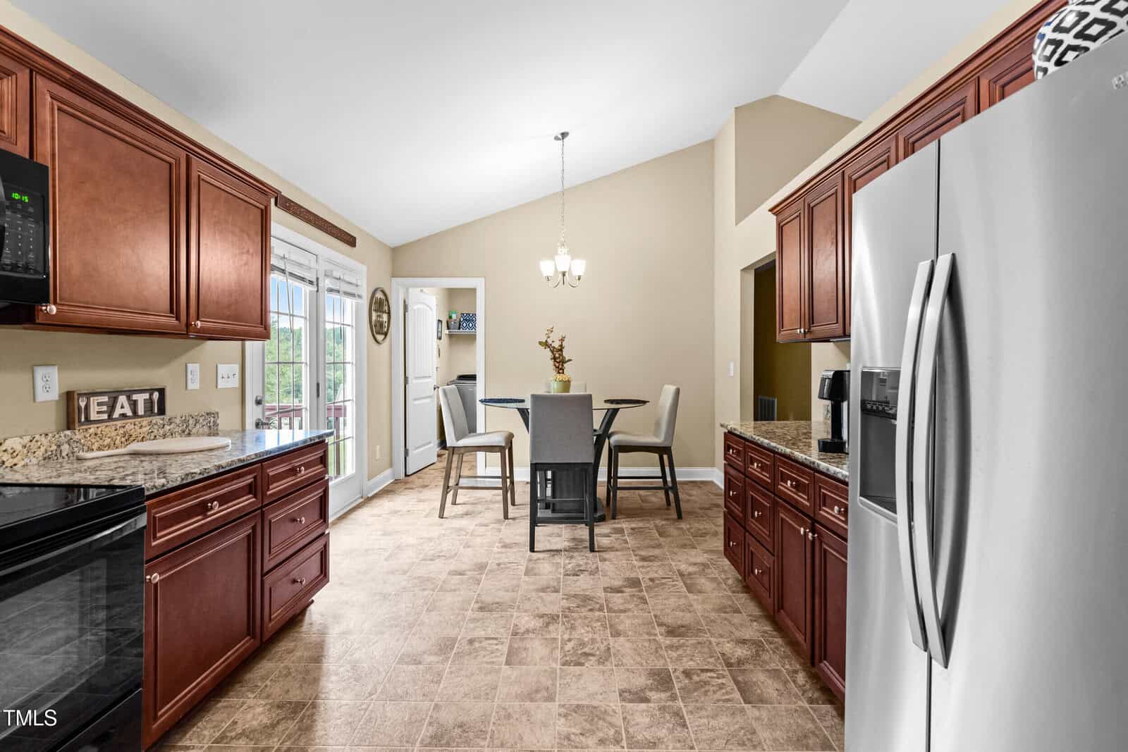 A kitchen with brown cabinets, granite countertops, and stainless steel appliances opens into a dining area with a round table and four chairs. A chandelier hangs from the ceiling, and French doors lead outside.