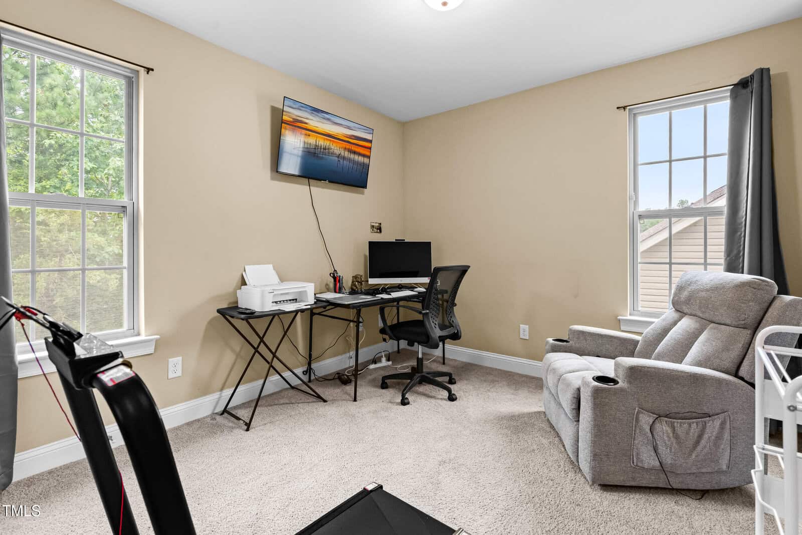 A home office with a desk holding a computer, printer, and office supplies. There is a black office chair, a gray recliner, two windows with curtains, beige walls, and a wall-mounted TV displaying a sunset.