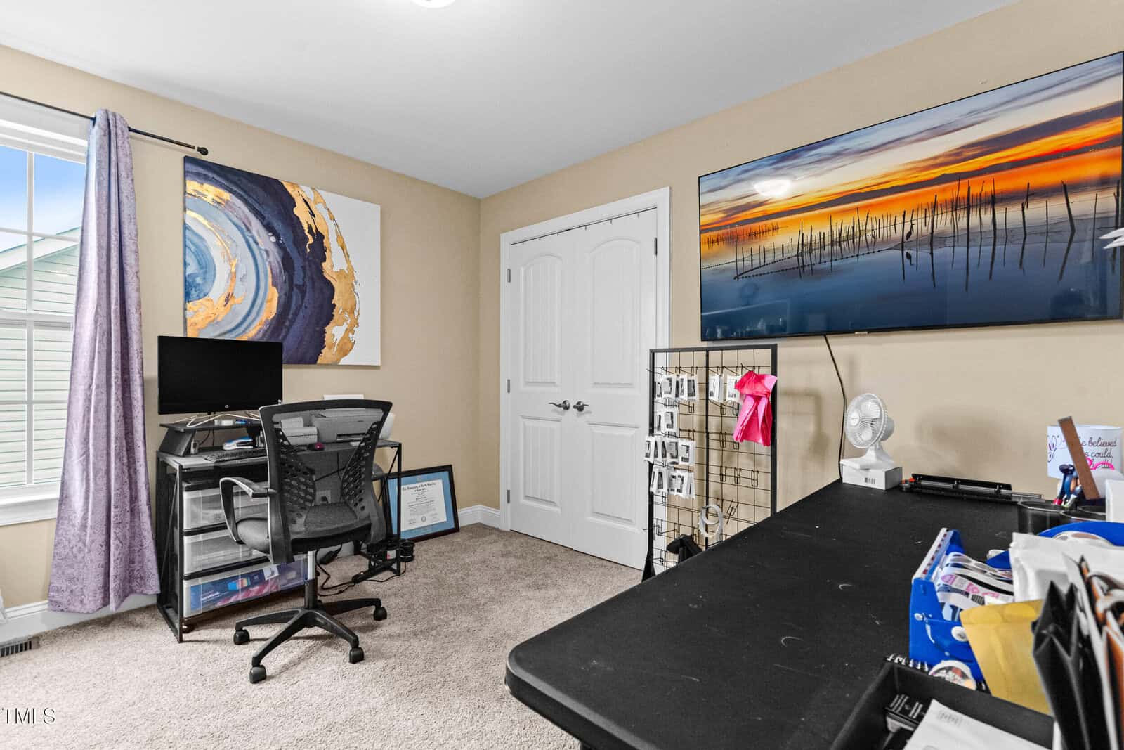 Home office with desk, office chair, computer monitor, wall art, TV displaying a sunset, a wire grid organizer with ribbons, and various items on a black table. Light enters through a window with purple curtains.