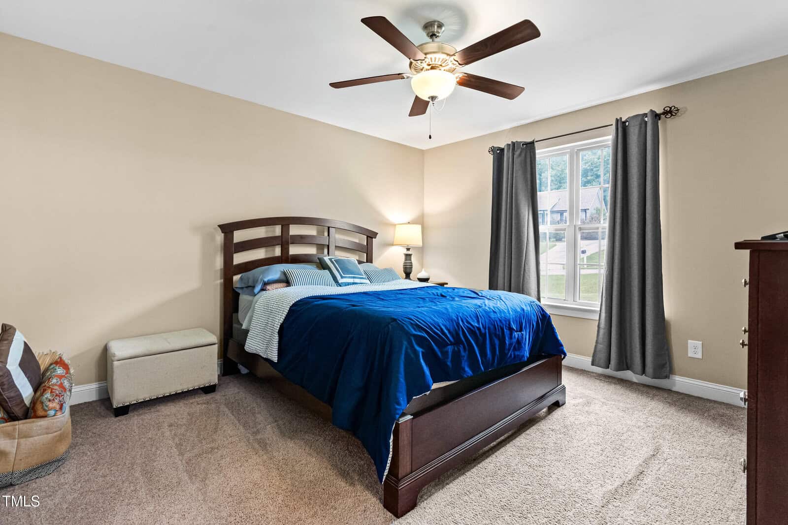 Bedroom with a wooden bed frame, blue bedding, a side table with a lamp, a ceiling fan, a window with gray curtains, beige walls, and carpeted floor.