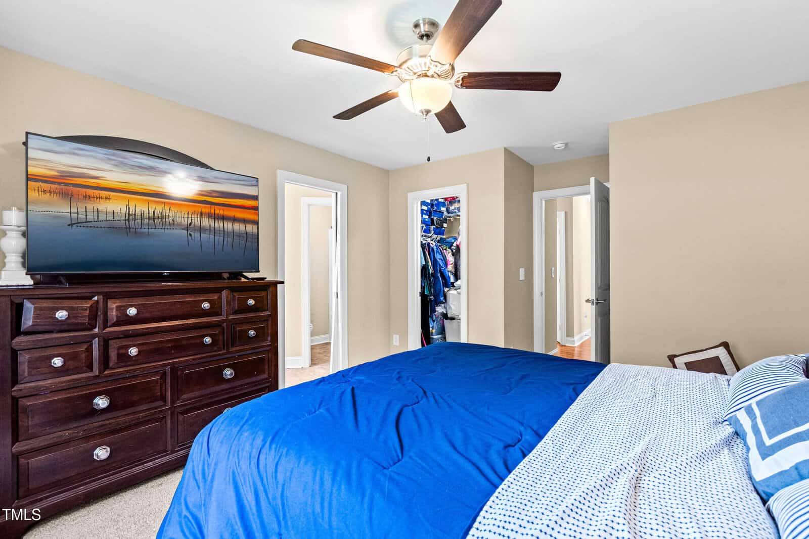 A bedroom with beige walls, a ceiling fan, a large dresser with a flat-screen TV, a bed with blue and white bedding, and open doors revealing a closet and a hallway.