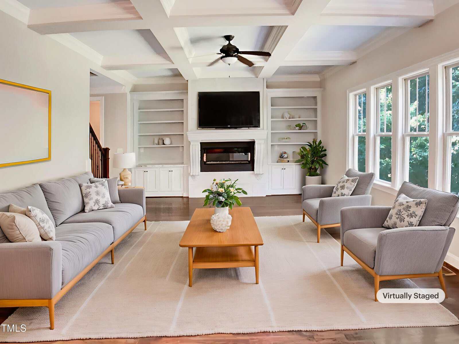 Bright living room with light gray sofa and armchairs, wooden coffee table, built-in shelves around a fireplace with a TV above it, large windows on the right, and a ceiling fan. The space is virtually staged.