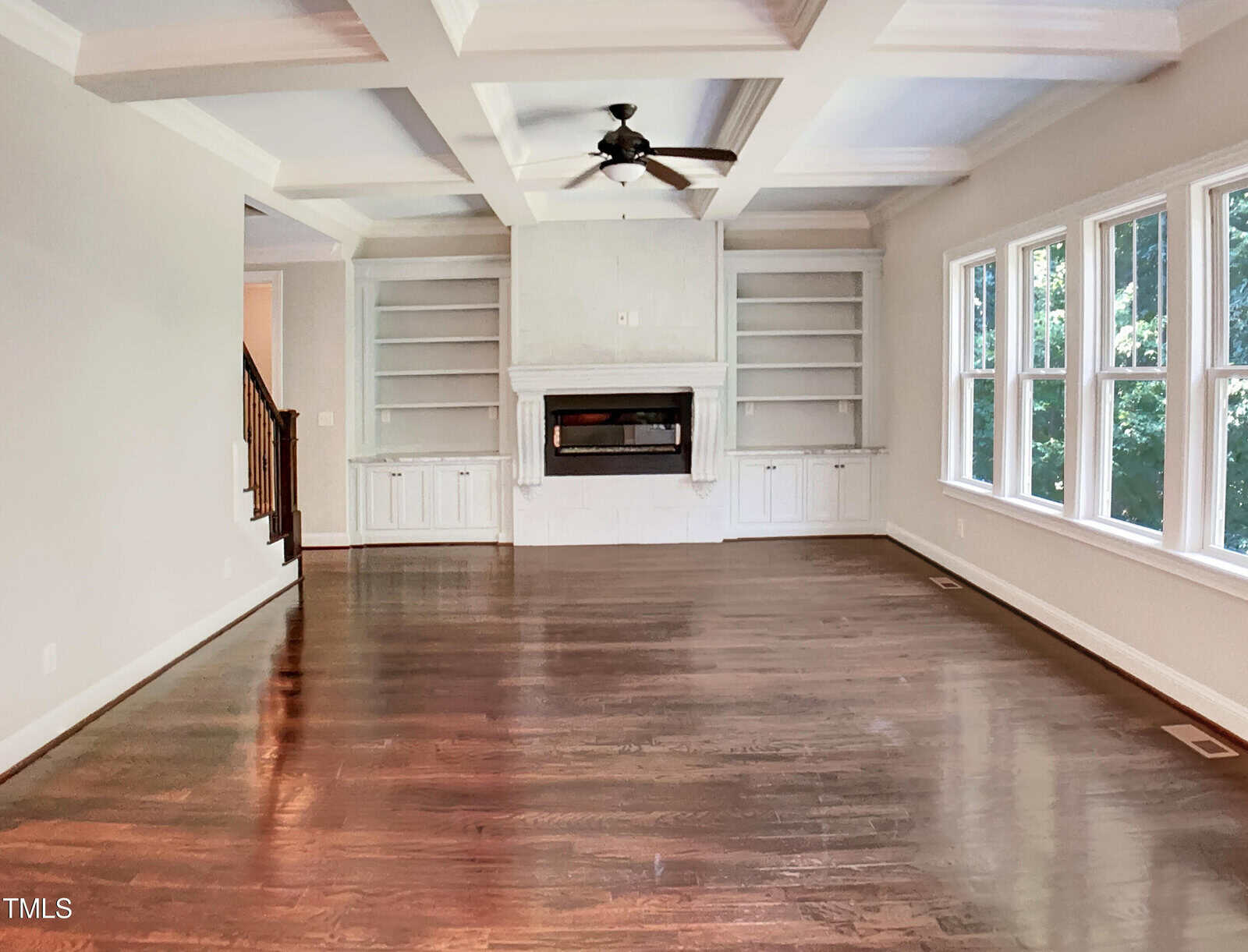 Empty living room with dark hardwood floors, white built-in shelves flanking a fireplace, coffered ceiling with a ceiling fan, and large windows along one wall letting in natural light.