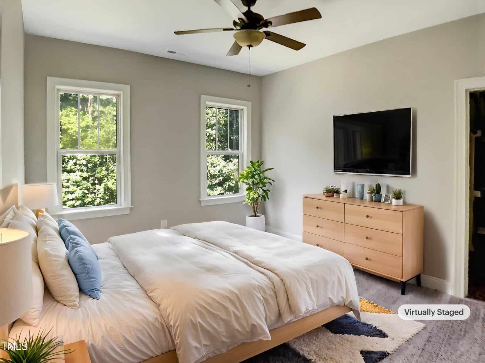 A modern bedroom with a large bed, wooden dresser, wall-mounted TV, two windows, a plant, and a ceiling fan. The room has light-colored walls and is bright with natural light. A "Virtually Staged" label appears in the corner.