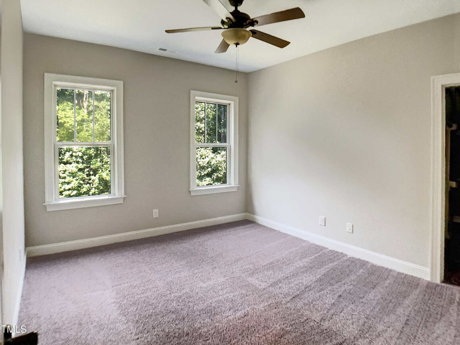 Empty room with beige carpet, light gray walls, two windows showing greenery outside, a ceiling fan with lights, and a small doorway visible to the right. Natural light fills the space.