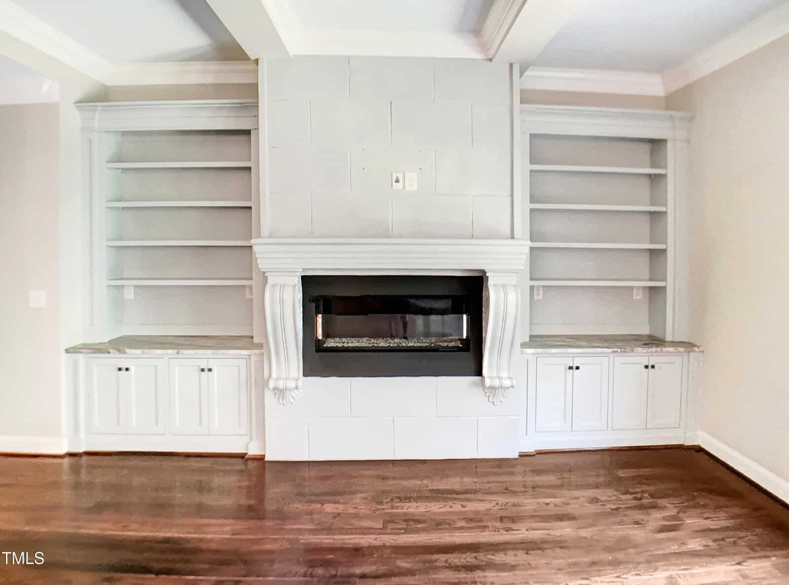 A white fireplace with a dark glass insert is centered between two built-in shelving units with cabinets below. The room has light walls, crown molding, and dark hardwood floors.