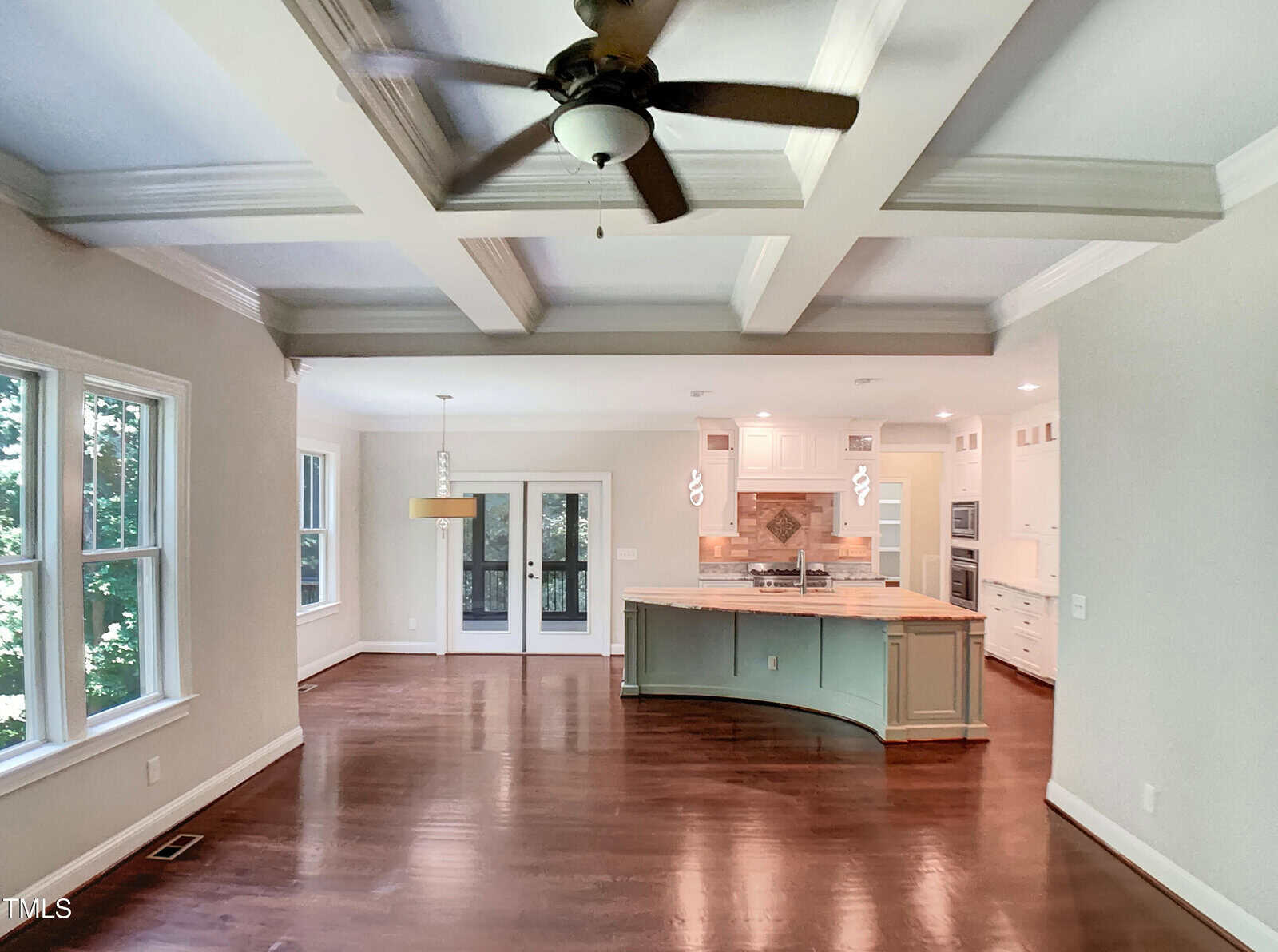 A spacious open-plan room with wood floors, a coffered ceiling with a ceiling fan, large windows, a kitchen with an island and pendant lights, and a dining area near glass doors leading outside.