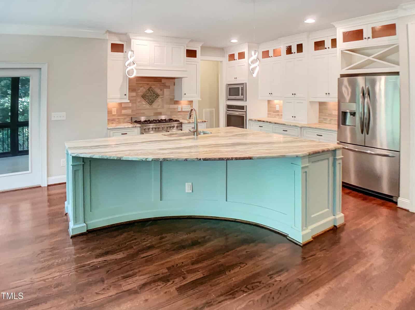 A modern kitchen with a large, light blue curved island, marble countertop, stainless steel appliances, white cabinetry, wood floors, and pendant lights.