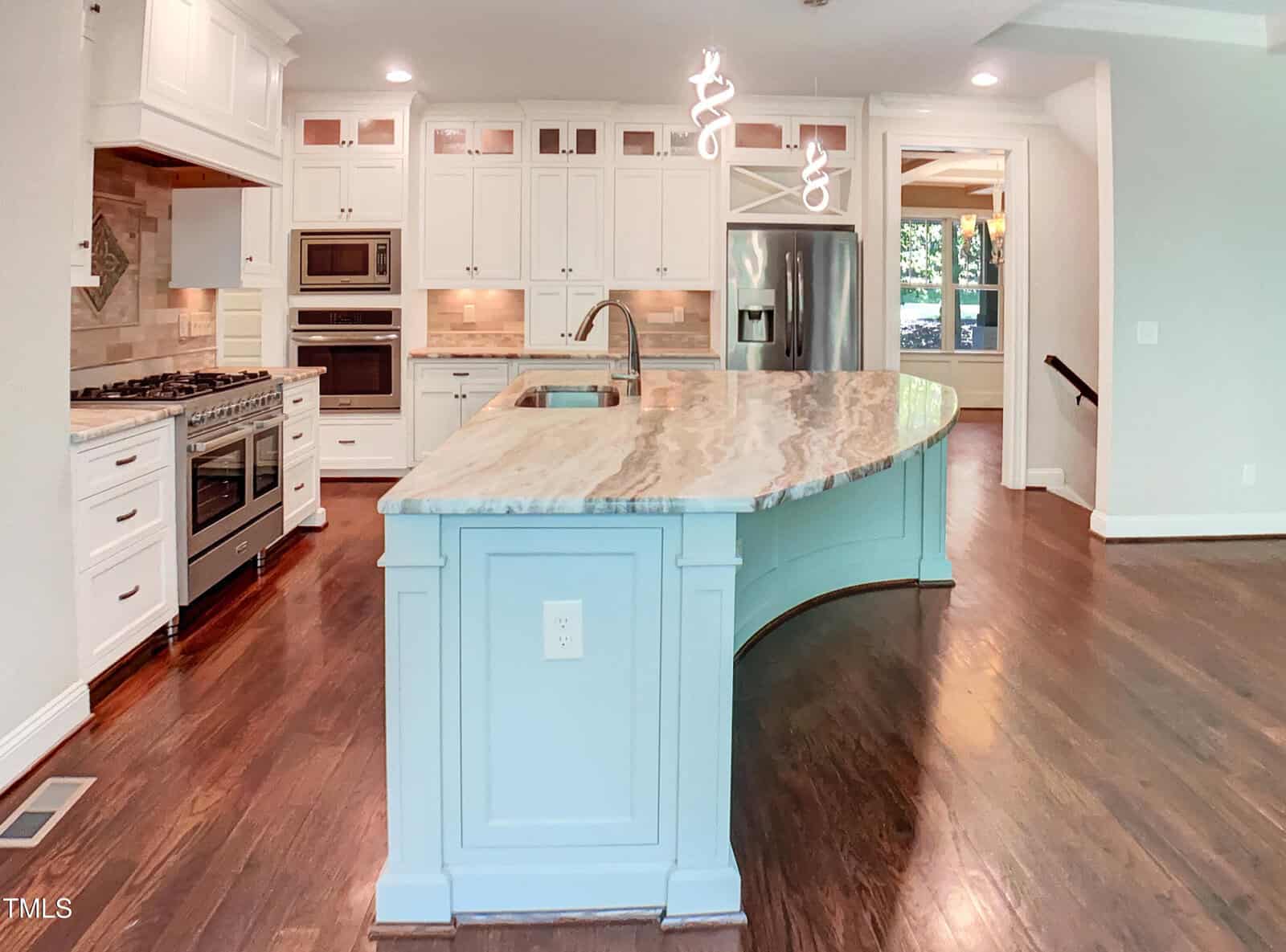 A modern kitchen with a large, curved marble island featuring a sink, white cabinets, stainless steel appliances, built-in oven and microwave, and dark hardwood floors. Two pendant lights hang above the island.