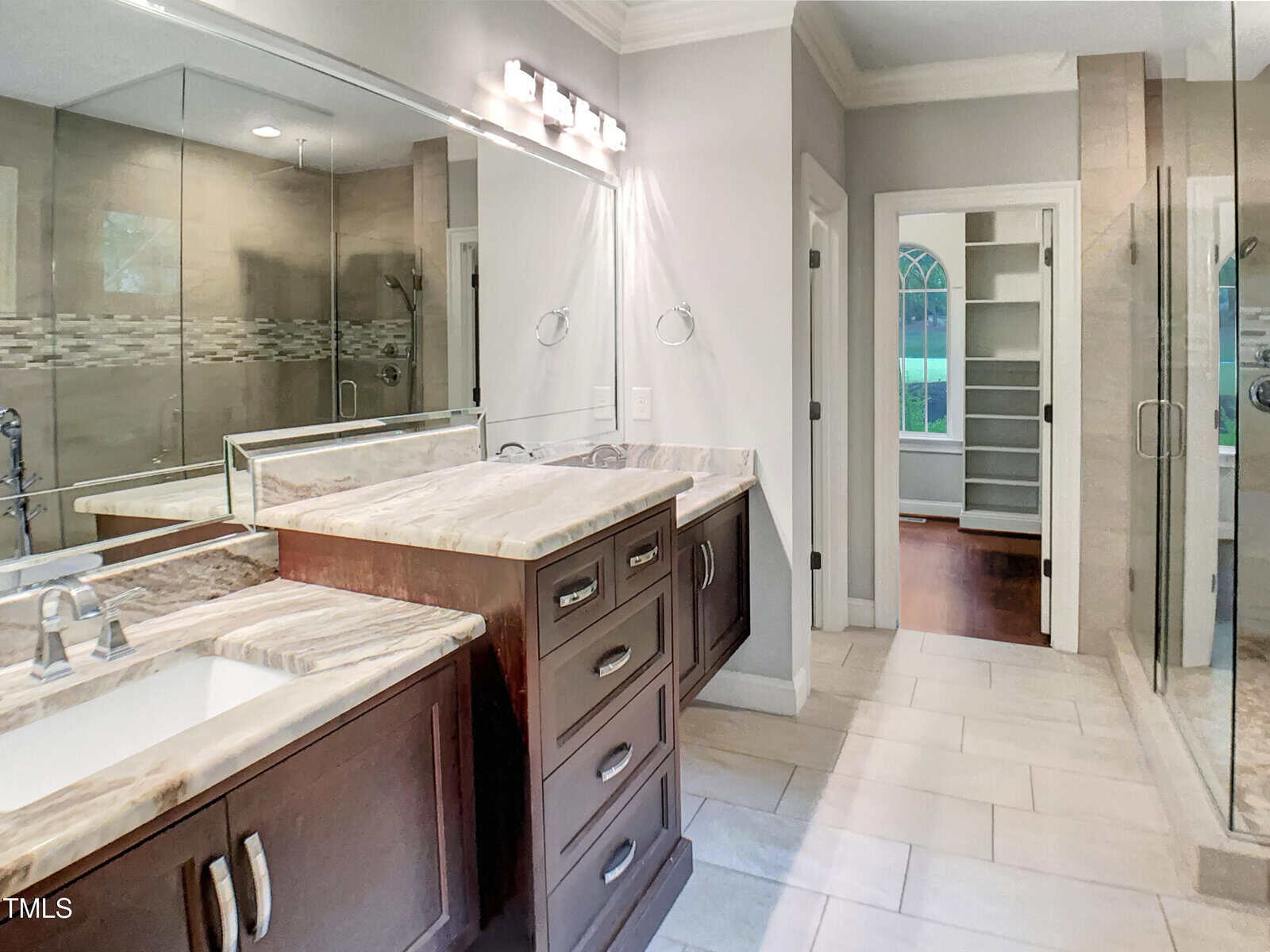 Modern bathroom with dual sinks, dark wood cabinets, marble countertops, glass-enclosed shower, light tile floor, large mirror with overhead lighting, and an open doorway leading to a walk-in closet with shelves.