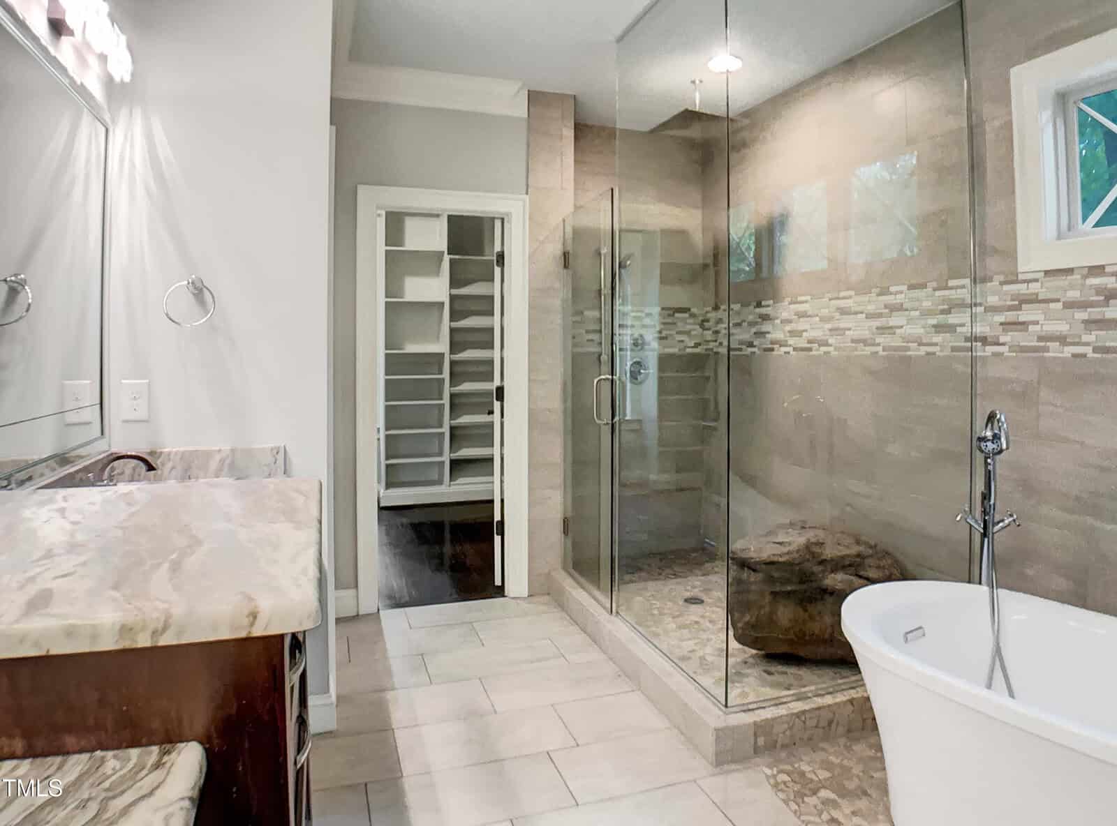 Modern bathroom with a glass shower, freestanding tub, marble countertop, and double sinks. Light tile flooring and walls, with an open doorway leading to a walk-in closet with built-in shelving.