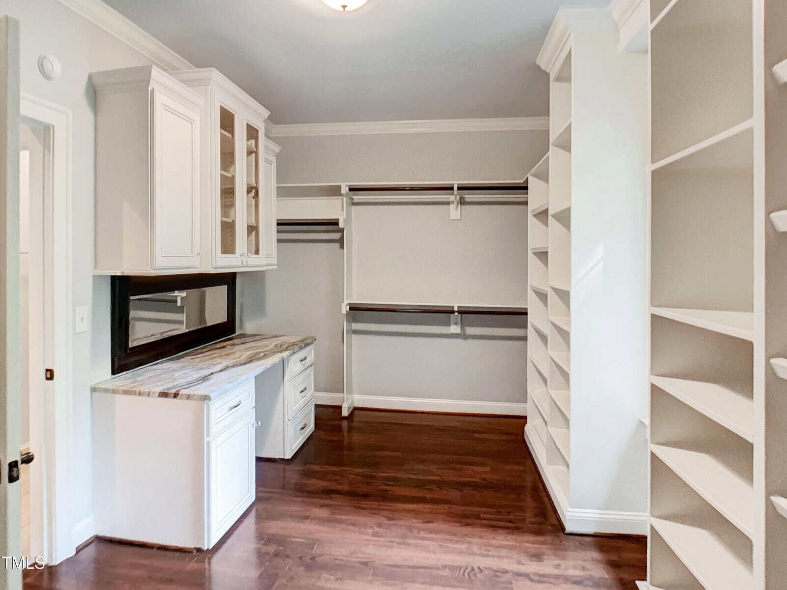 Spacious walk-in closet with white shelves, hanging rods, a marble-topped desk, cabinets with glass doors, and dark hardwood flooring under bright lighting.