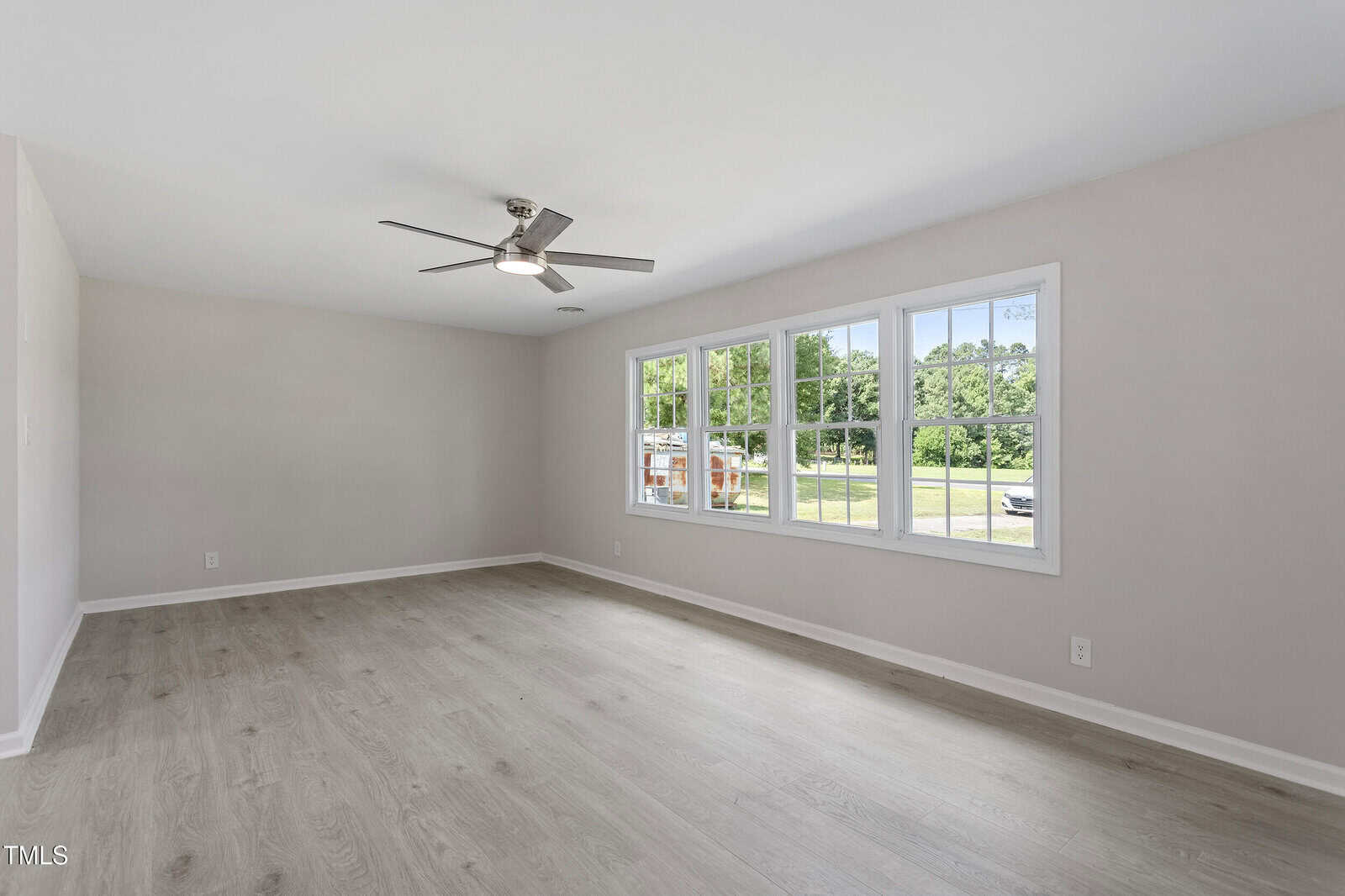 A large, empty room with light-colored wood flooring, white walls, a ceiling fan with lights, and three large windows letting in natural light. Trees and a parked vehicle are visible outside.