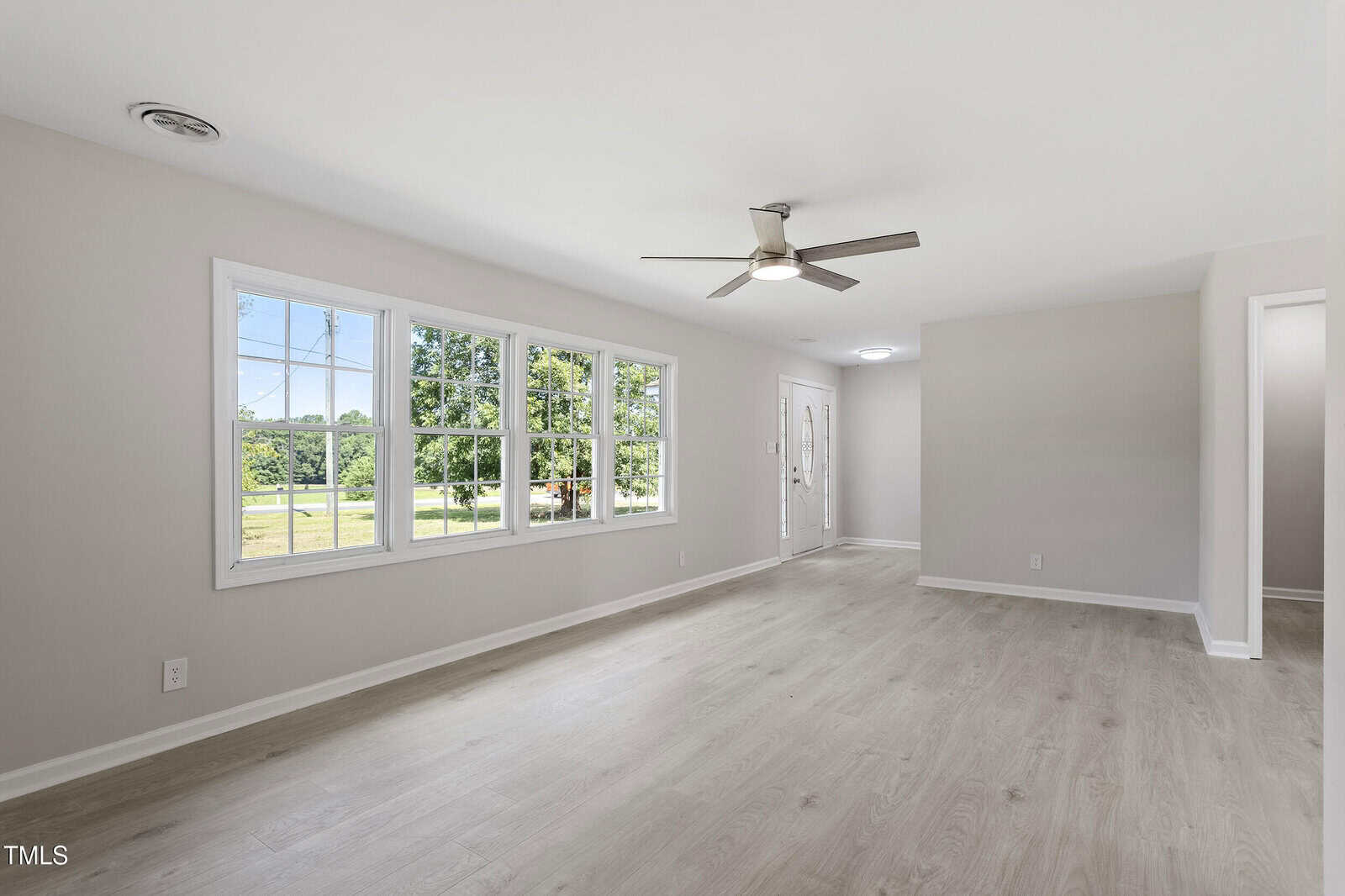 Spacious living room with light wood flooring, large windows showing a green outdoor view, white walls, and a ceiling fan with light. A doorway and main entrance are visible in the background.