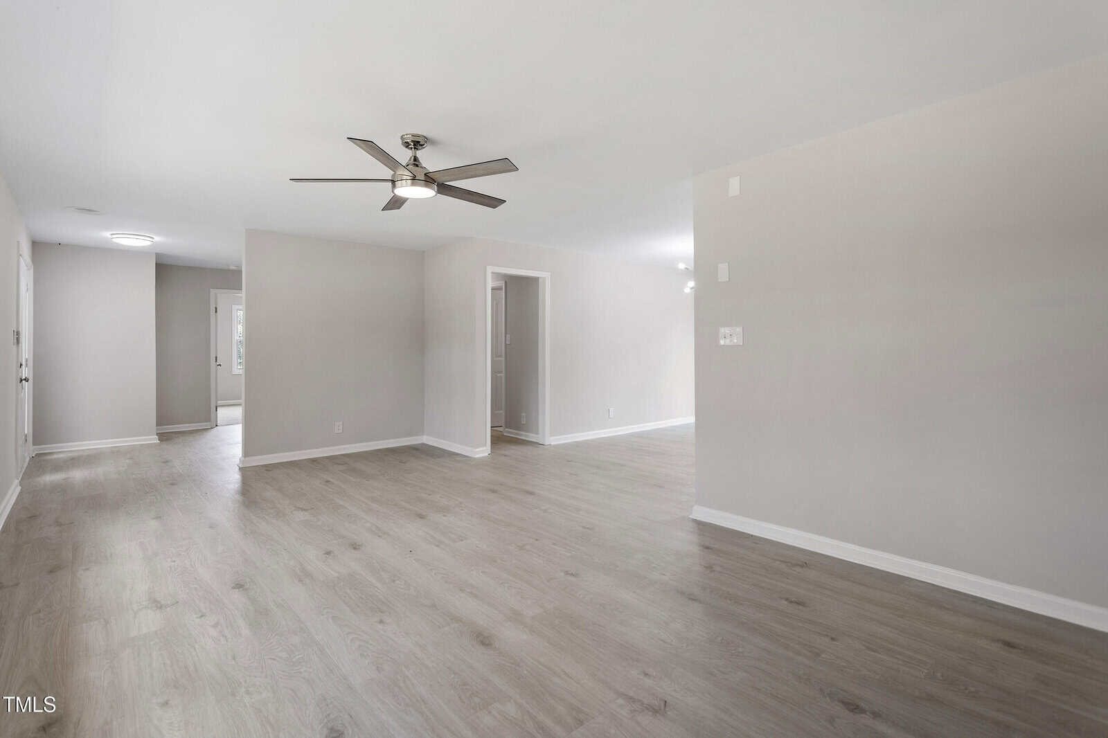 A spacious, empty living room with light wood flooring, beige walls, a ceiling fan with lights, and open doorways leading to other rooms in a modern home. Natural light enters from windows in the background.