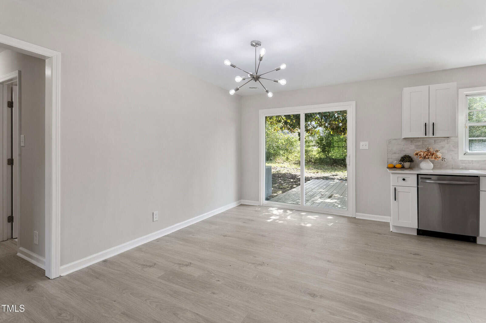 A bright, empty dining area with light wood floors, a modern chandelier, and sliding glass doors leading to an outdoor deck. The kitchen with white cabinets and stainless steel appliances is partially visible on the right.