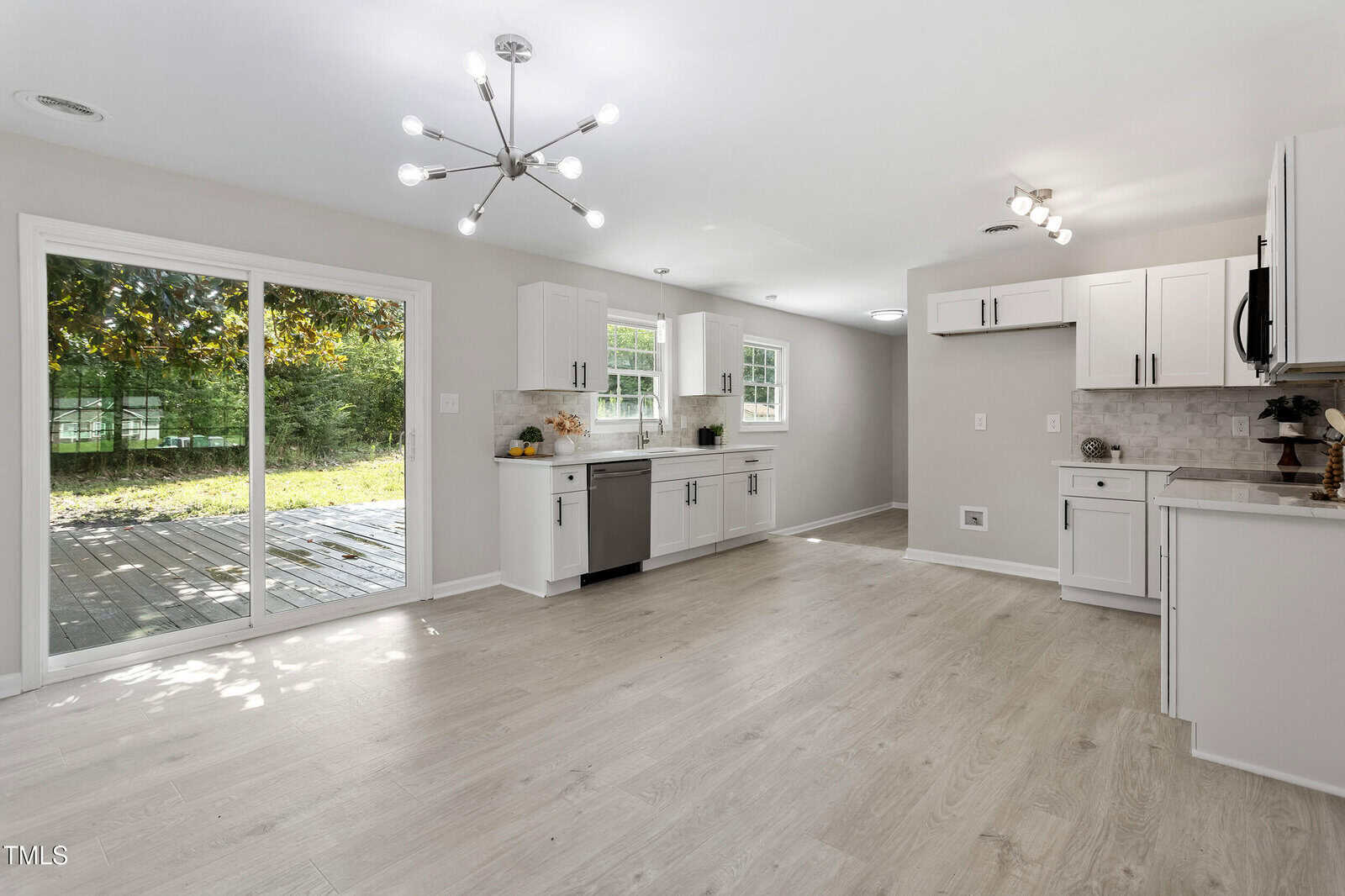 Bright, modern kitchen with white cabinets, light wood flooring, stainless steel appliances, and a sliding glass door opening to a backyard deck. Large windows let in natural light. Contemporary light fixtures are on the ceiling.
