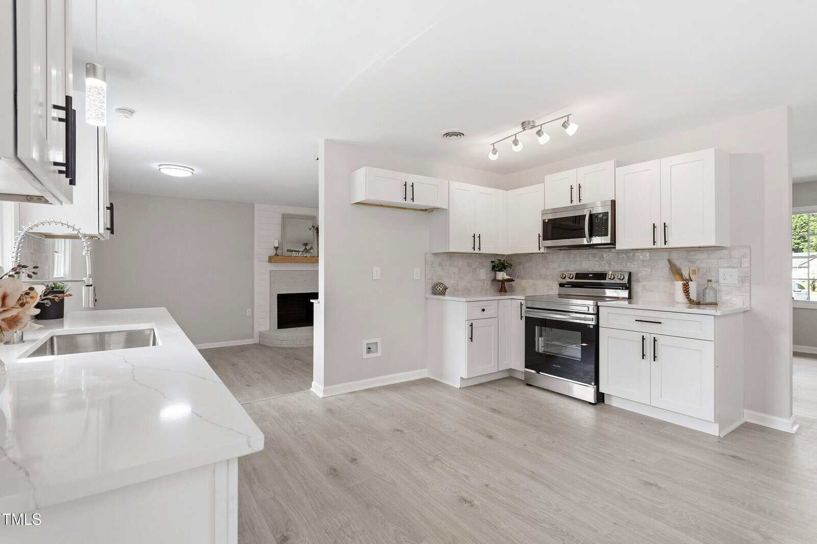 A modern kitchen features white cabinets, stainless steel appliances, light wood flooring, a white quartz countertop, and a partial view of a living area with a fireplace in the background.