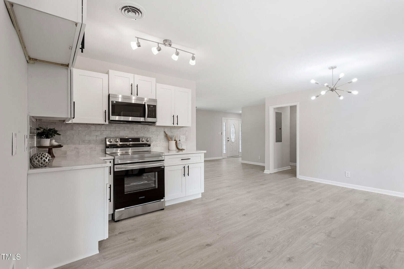 A modern, open-concept kitchen and living area with white cabinets, stainless steel appliances, light wood flooring, neutral walls, and contemporary lighting fixtures. The space appears clean and bright.