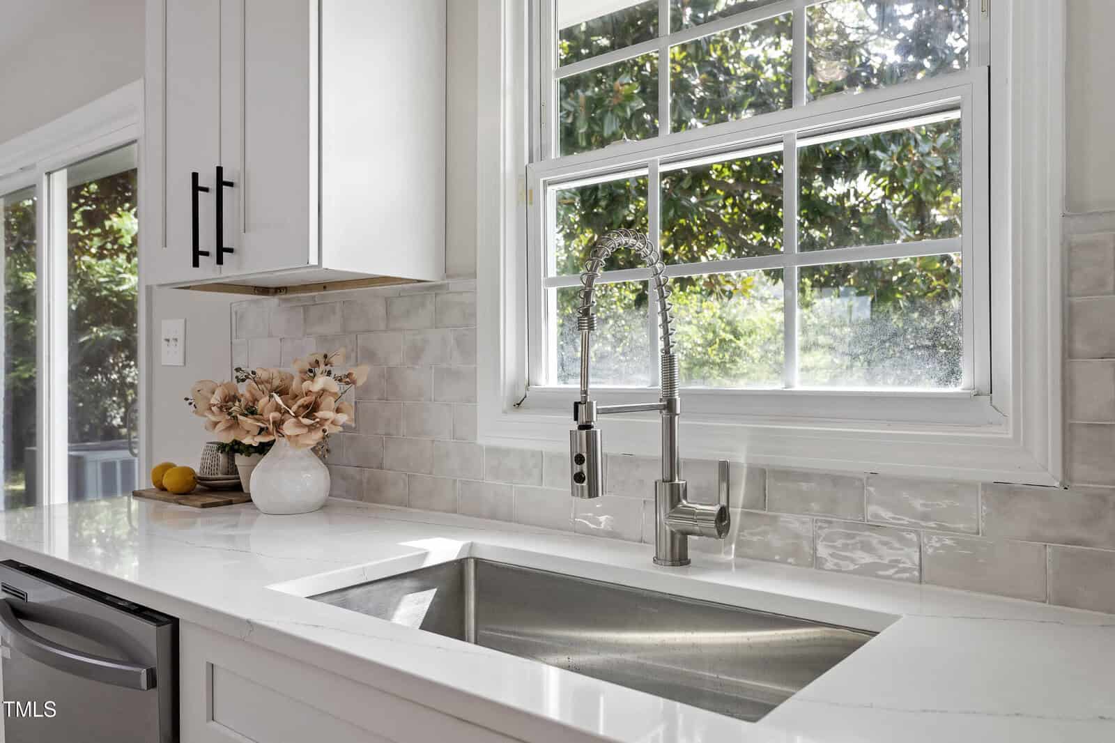 A modern kitchen sink with a stainless steel faucet beneath a window. The countertop is white with a vase of flowers and a lemon at the left. Light gray tile backsplash and white cabinetry are visible.