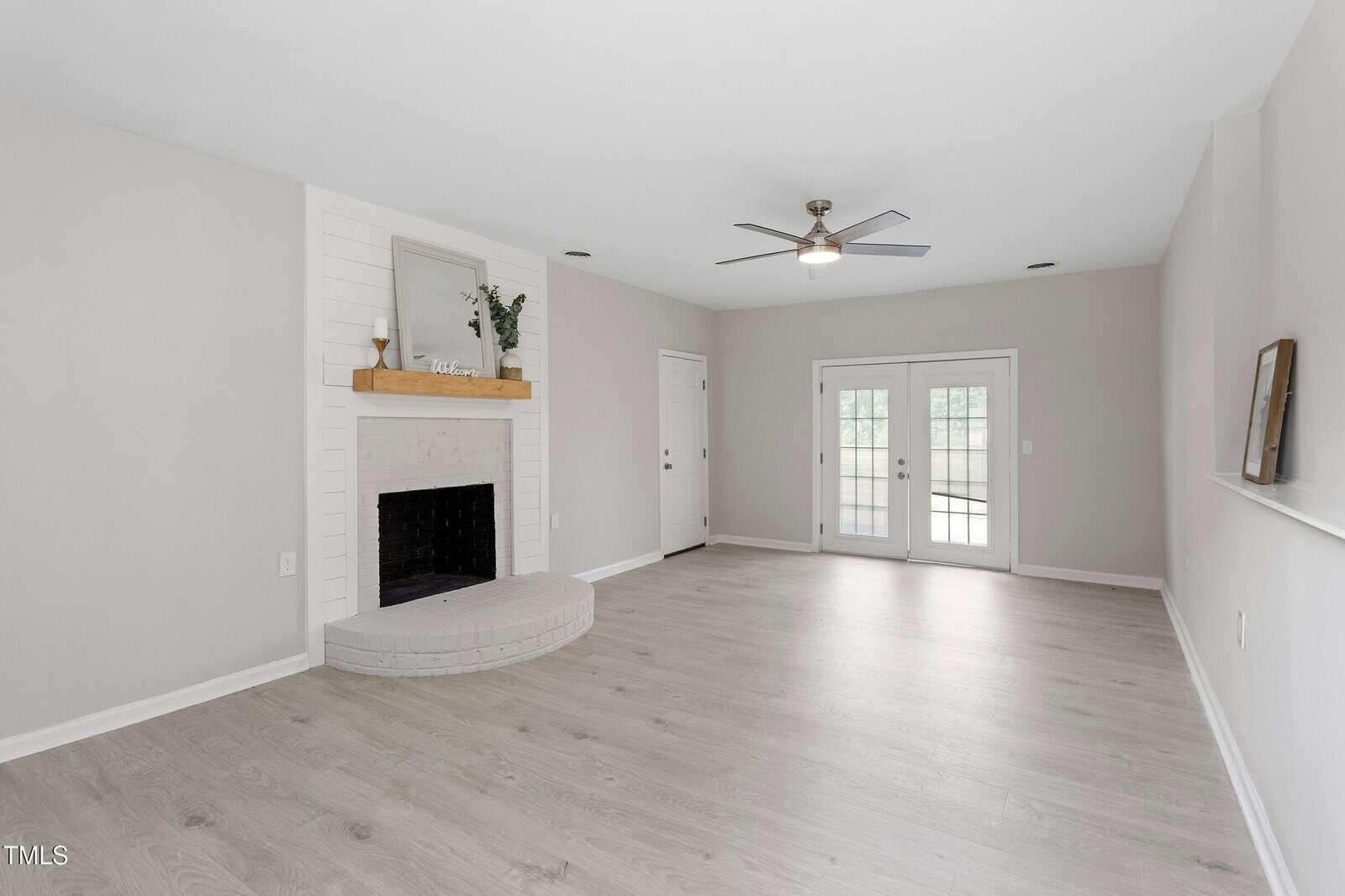 Bright living room with light wood floors, a white corner fireplace, ceiling fan, and double glass doors leading outside. Minimal decorations include a mirror, greenery, and a small framed sign on the mantel.