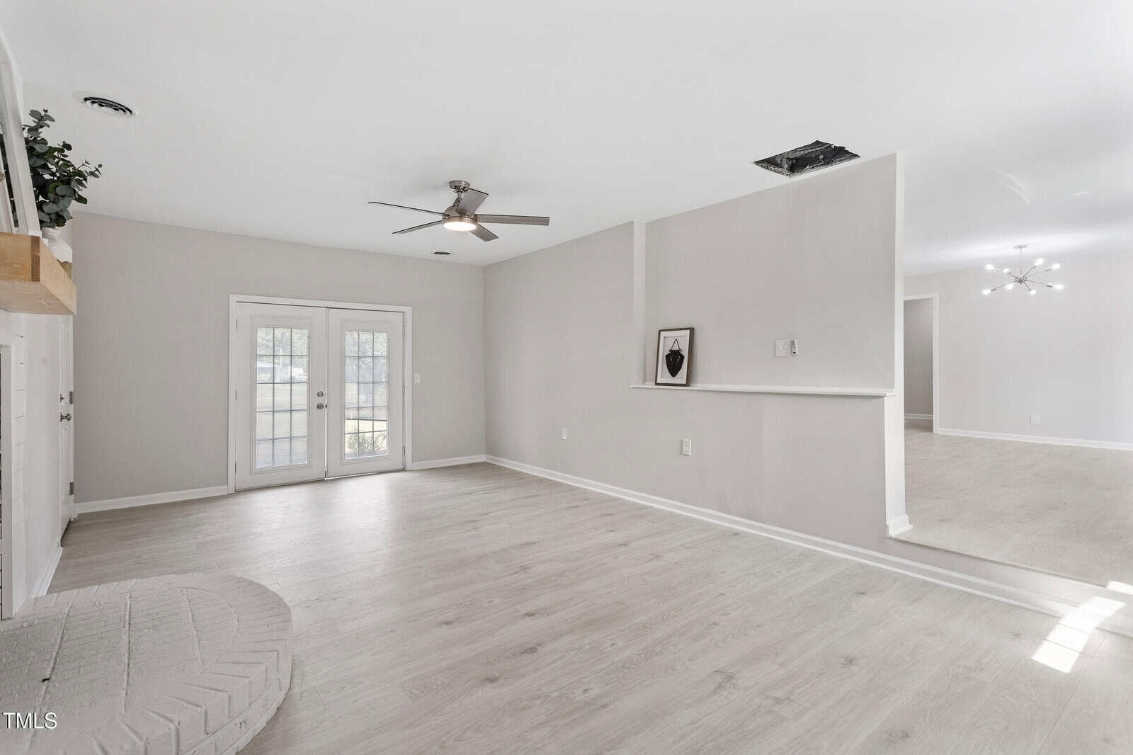 Bright, empty living room with light wood floors, white walls, a ceiling fan, French doors, and a small recessed shelf holding framed art. An open doorway leads to another room with a modern light fixture.