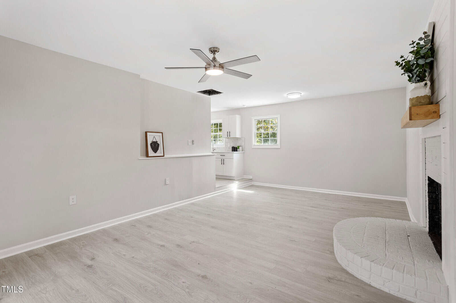 Bright, modern living room with light wood floors, white walls, a ceiling fan, a white brick fireplace, and an open view into a small kitchen with white cabinets and natural light from two windows.