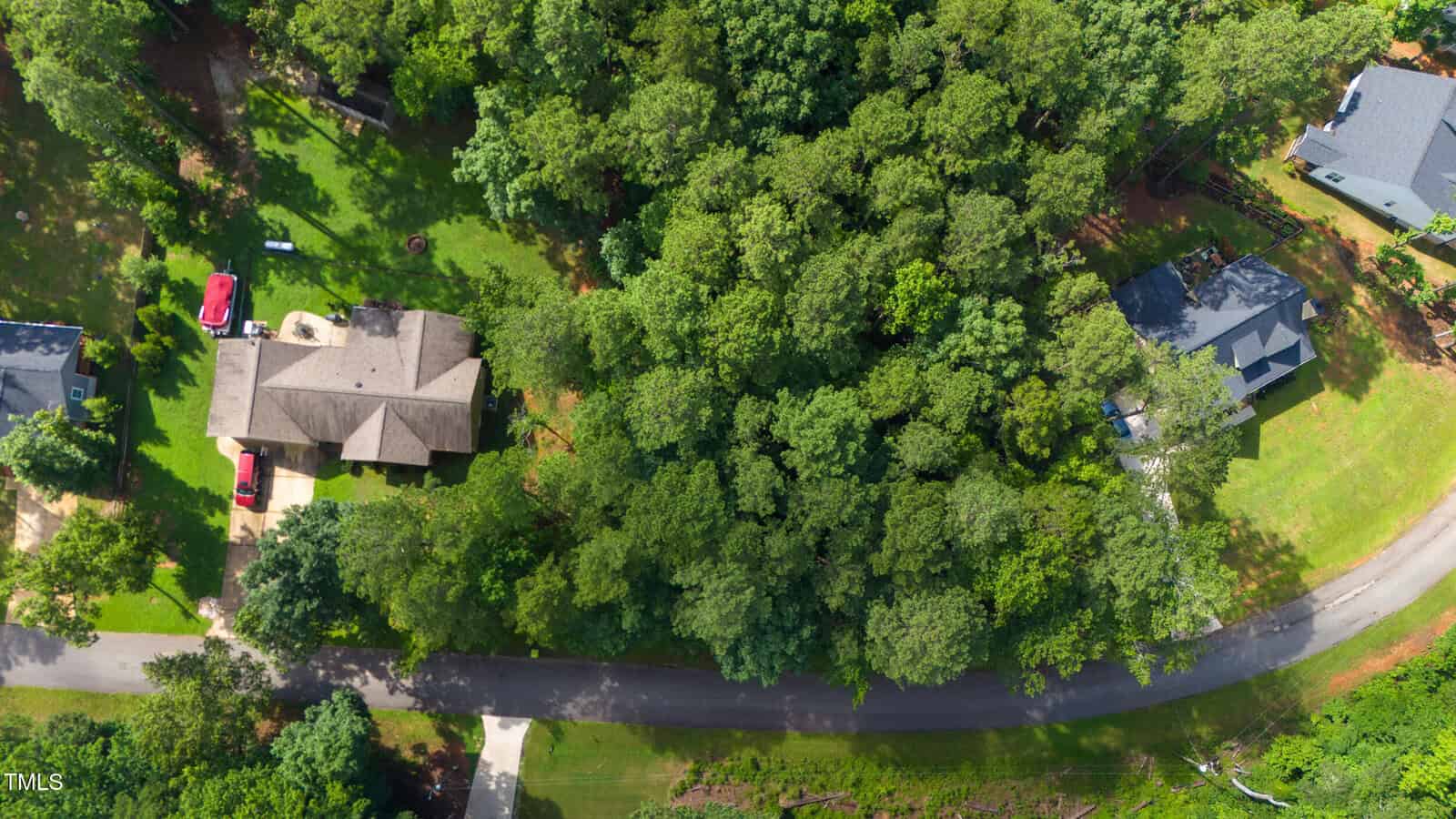 Aerial view of a suburban neighborhood with houses surrounded by dense green trees, a curved road, driveways, and a few parked vehicles. Forested area separates two homes.