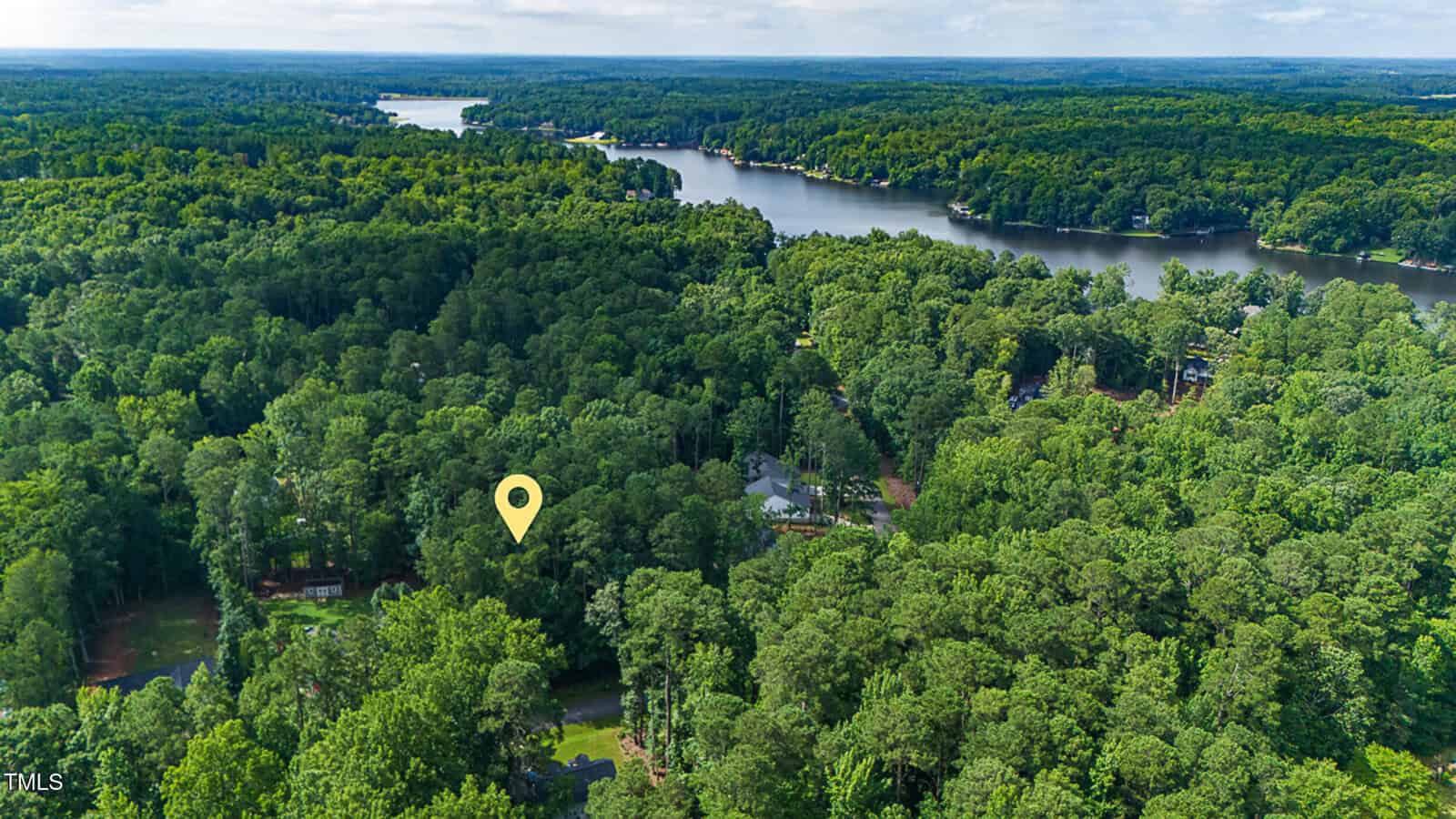 Aerial view of a forested lakeside community with dense green trees, winding roads, scattered houses, and a large lake. A yellow location marker highlights a specific spot among the trees near the water.