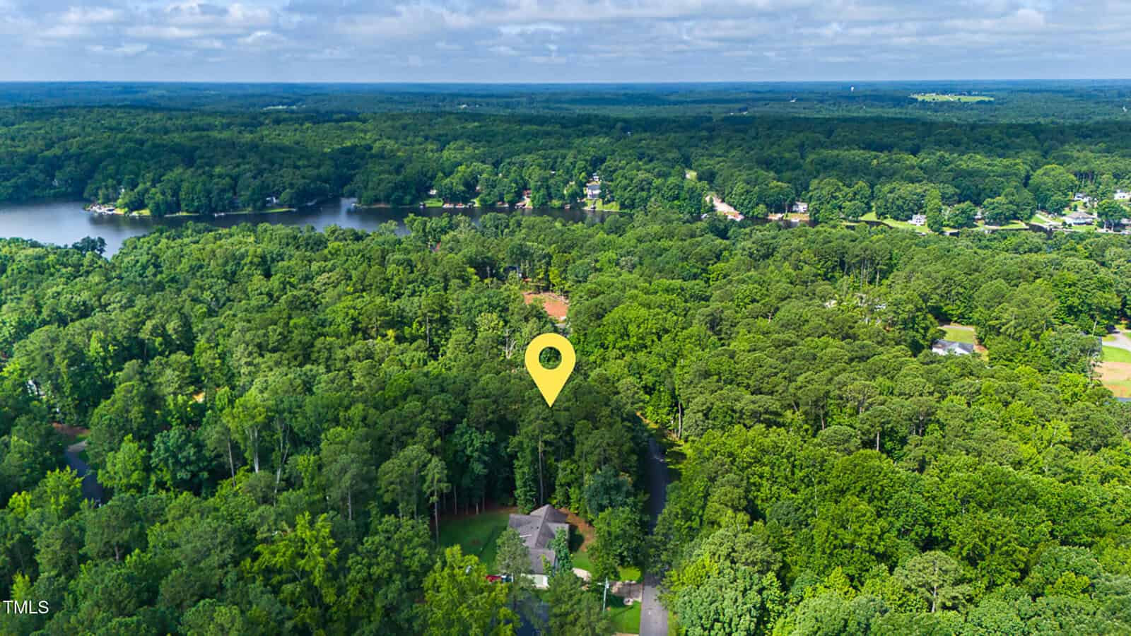 Aerial view of a wooded area with a body of water nearby. A yellow location marker is placed above a house surrounded by dense trees. Other homes and more water are visible in the background under a partly cloudy sky.