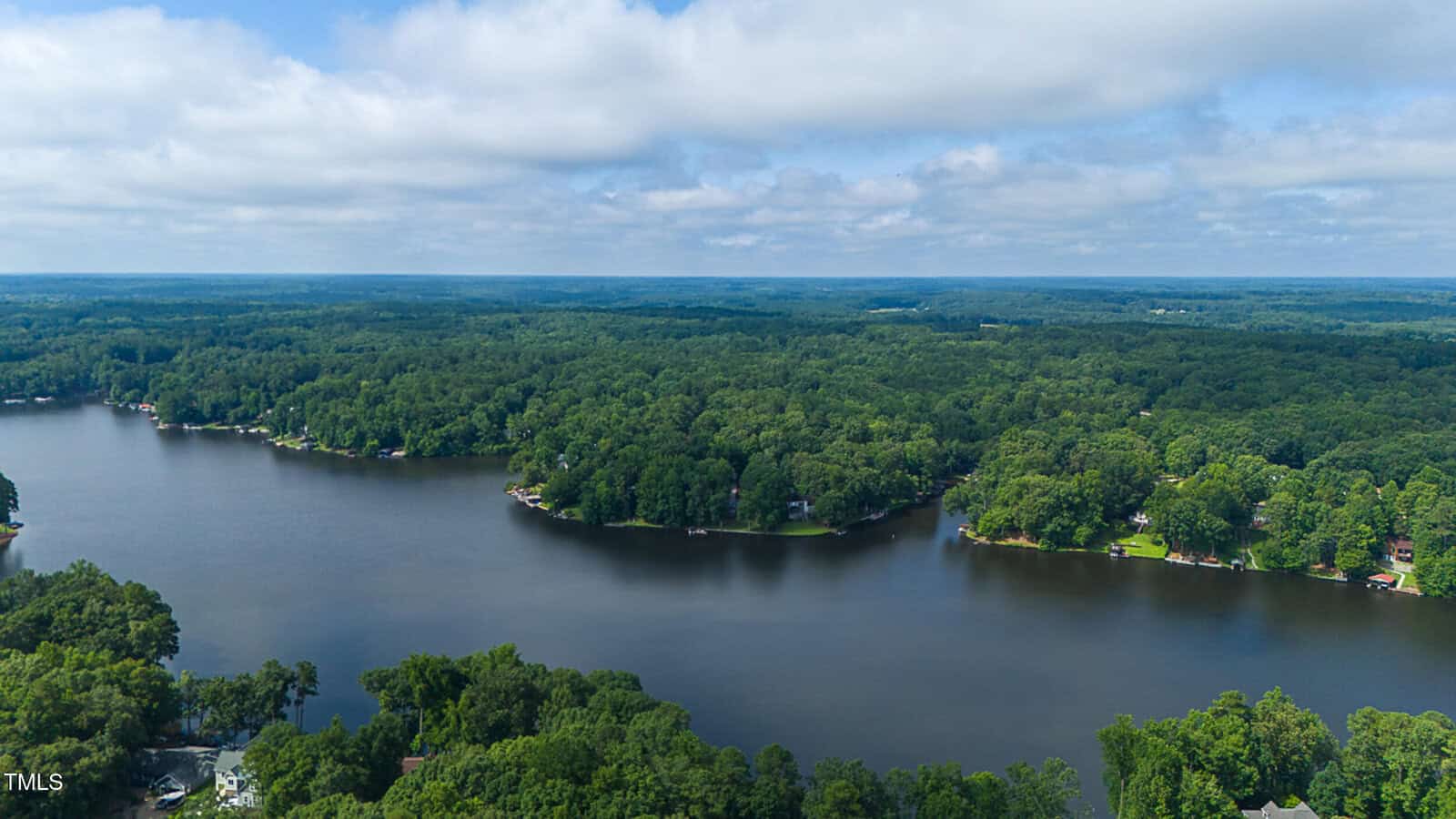 Aerial view of a large, calm lake surrounded by dense green forest and scattered homes along the shoreline under a partly cloudy sky.