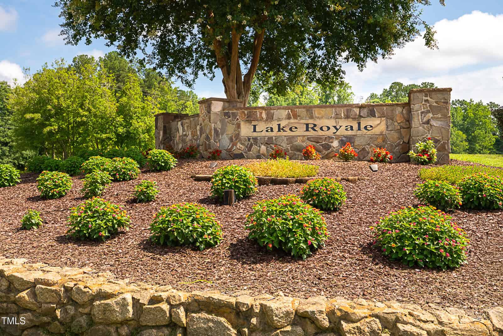 A stone sign reading “Lake Royale” stands behind landscaped shrubs and mulch, bordered by a low stone wall. A large tree and greenery are visible in the background under a partly cloudy sky.