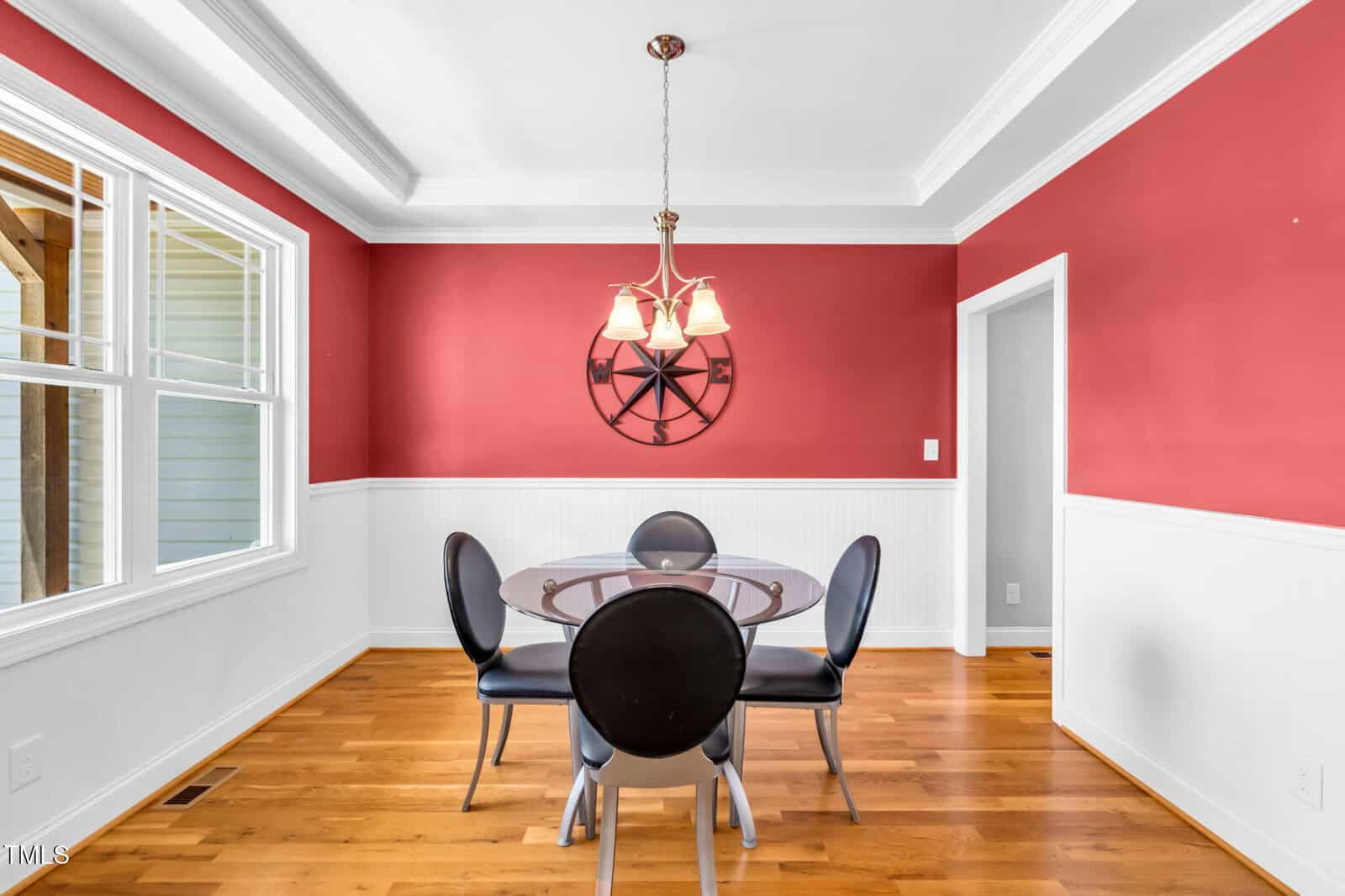 A dining room with red upper walls and white wainscoting, a glass-top round table with four black chairs, hardwood floors, a wall clock, and a ceiling light fixture. Large windows bring in natural light.